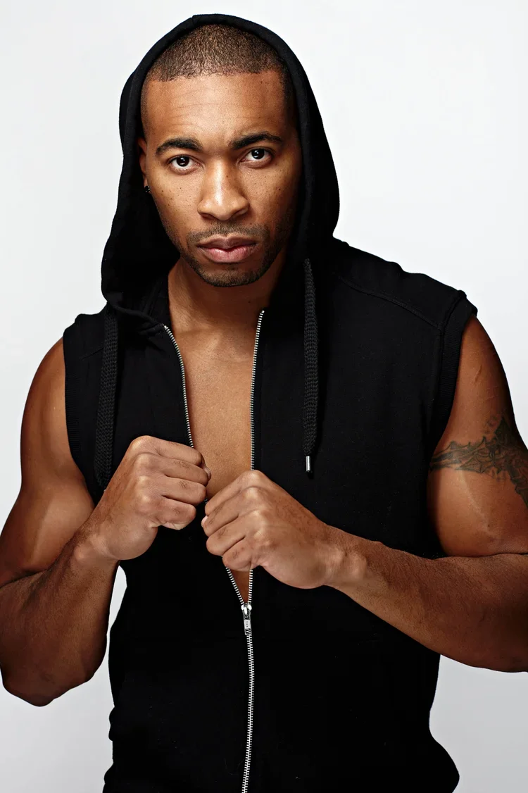 A muscular man in a black sleeveless hoodie with the hood up, standing against a plain white background, looking directly at the camera with a serious expression.