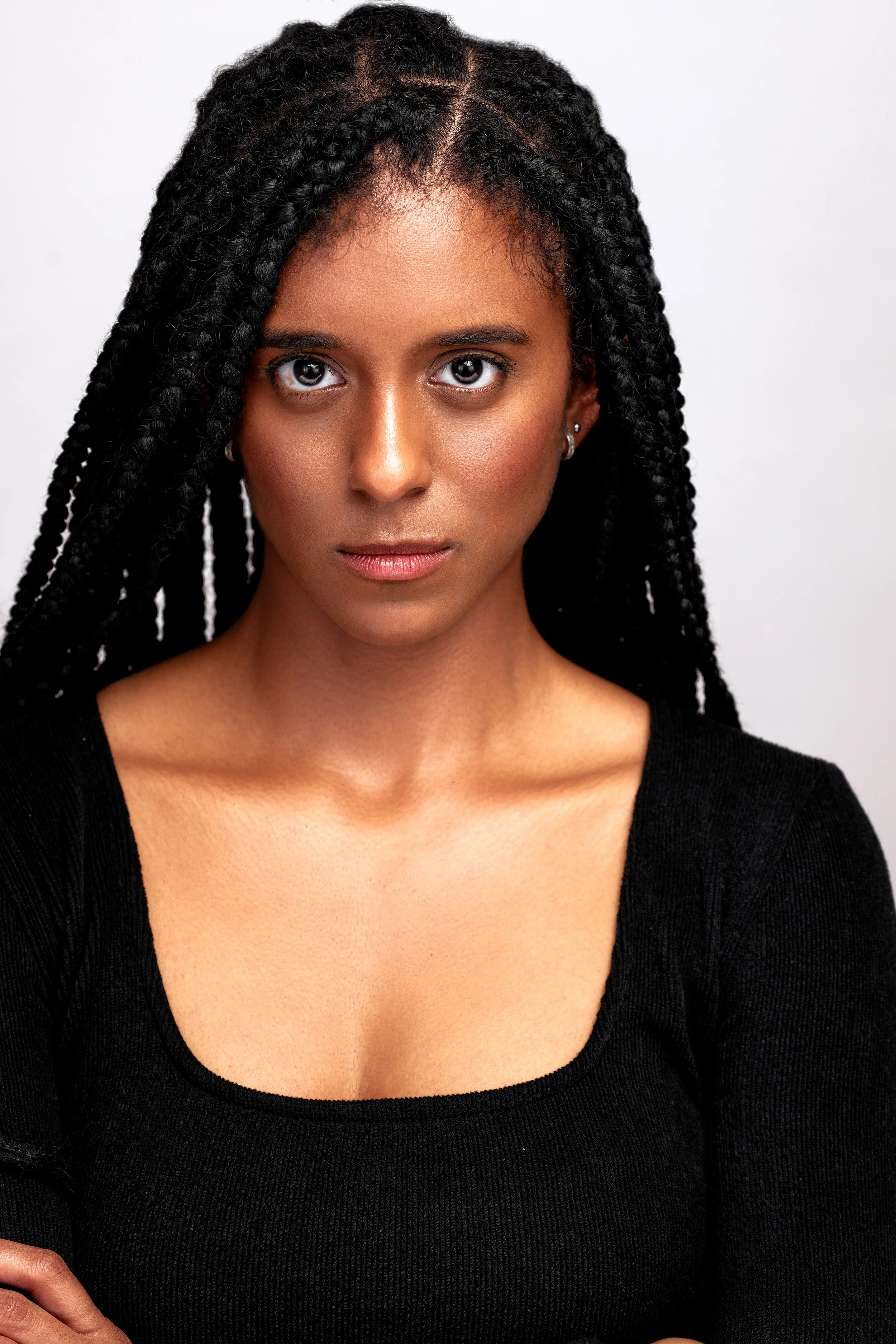 Close-up of a woman with medium-dark skin, long black braided hair, and wearing a black top, looking directly at the camera.