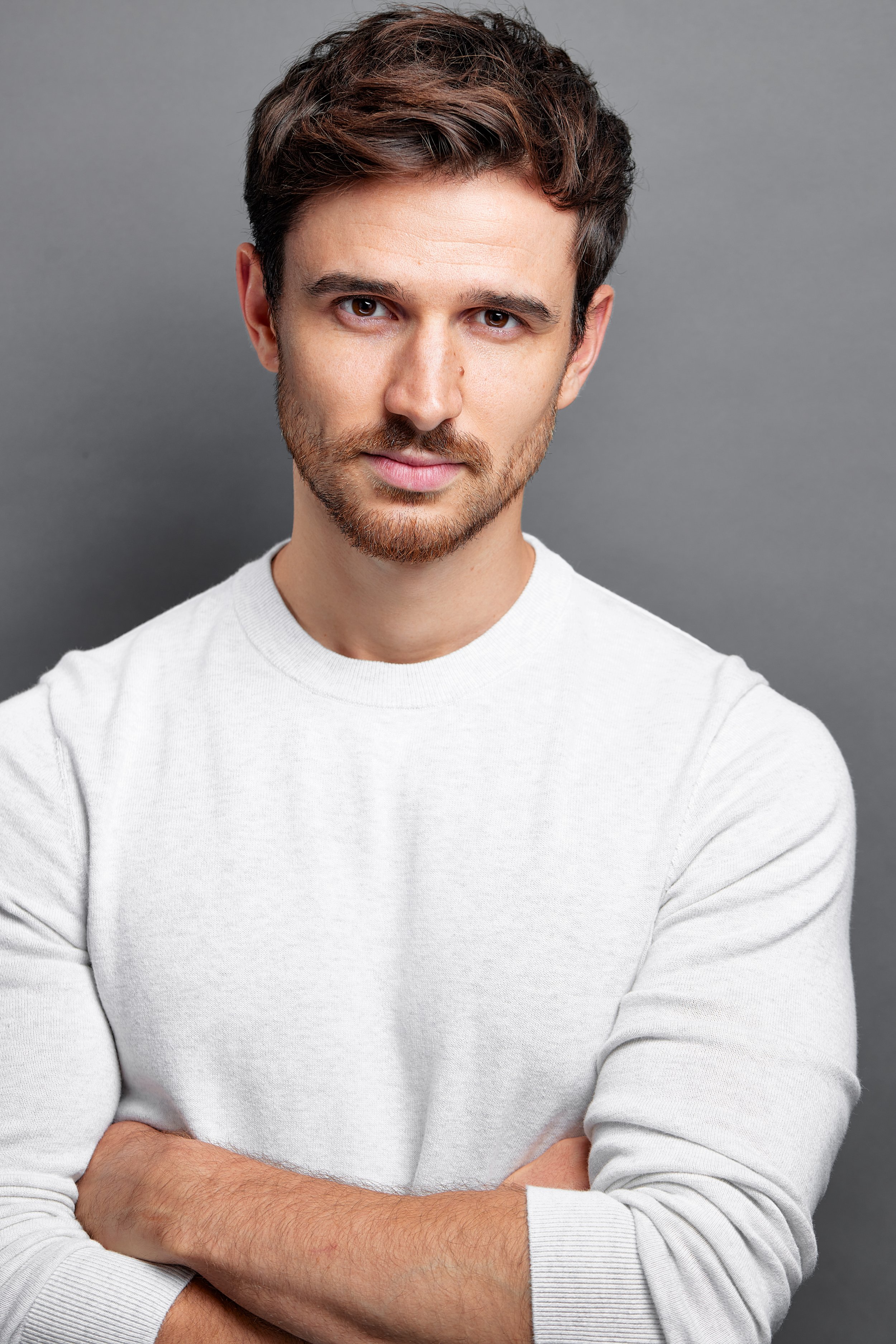 A young man with short brown hair, light skin, and facial hair, wearing a white sweatshirt with arms crossed, standing against a gray background.