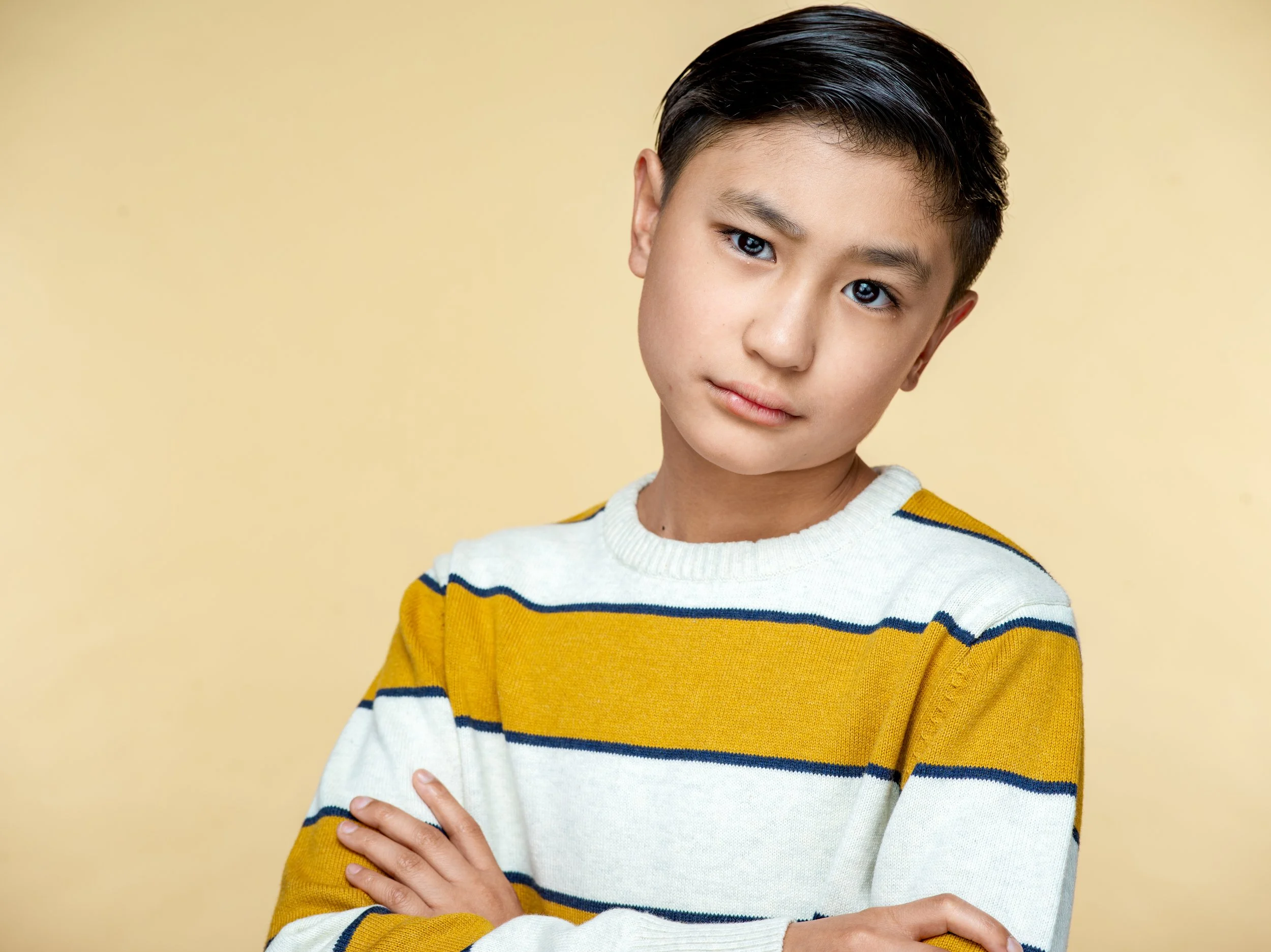 Young person with dark hair and light skin, wearing a yellow and white striped sweater, posing against a plain beige background.