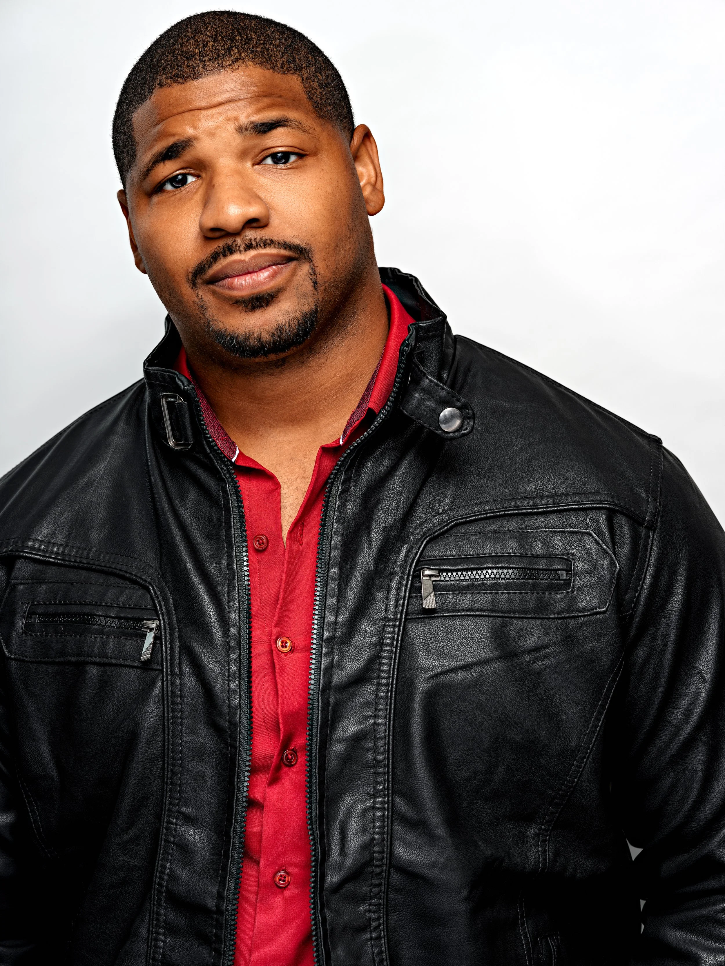 A young man with short hair, a beard, and a mustache, wearing a red shirt and a black leather jacket, looking at the camera with a slight smile against a plain white background.