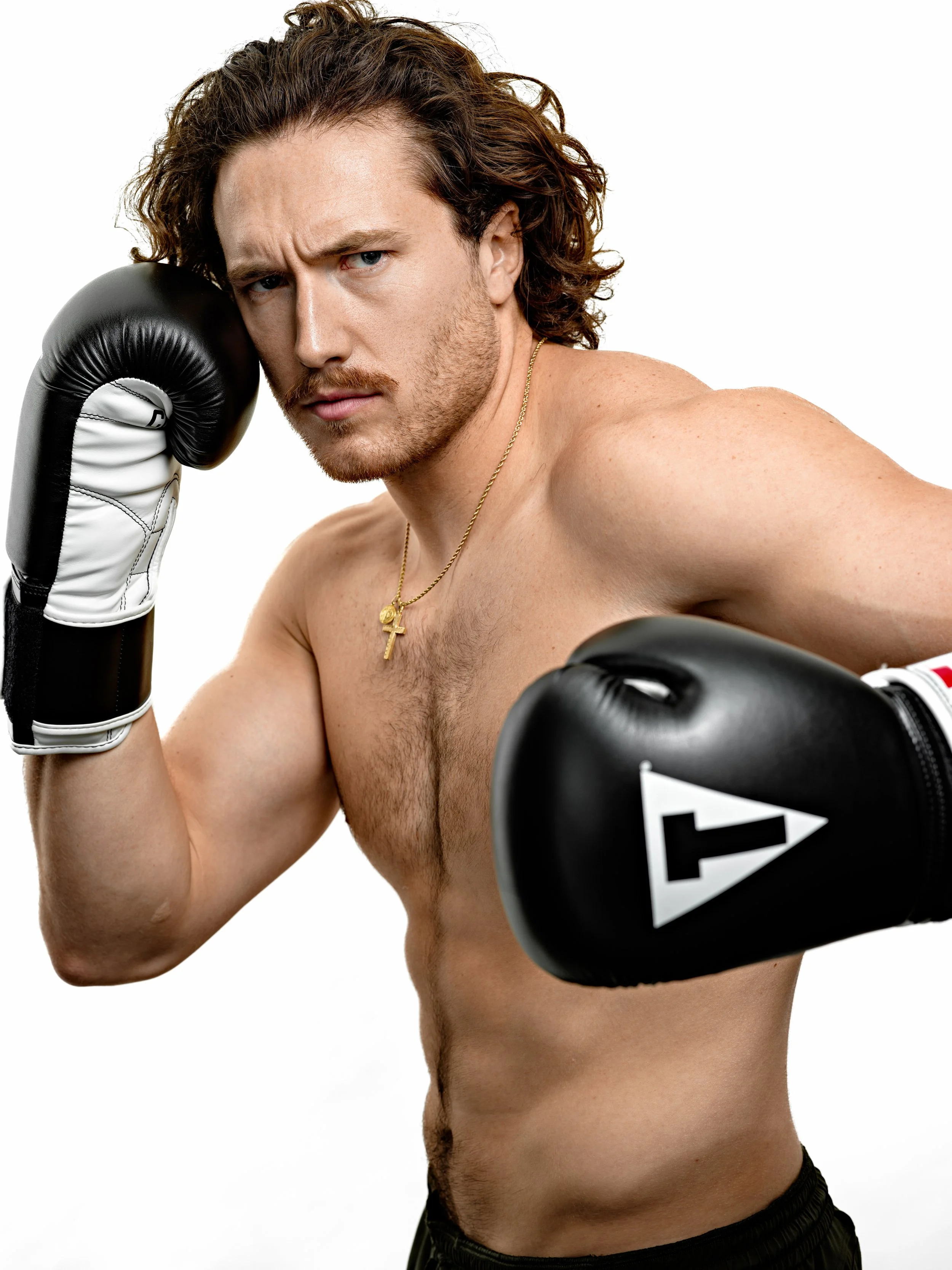 A shirtless man with curly brown hair, blue eyes, and a goatee poses with a boxing stance, wearing black boxing gloves and a gold cross necklace, against a white background.