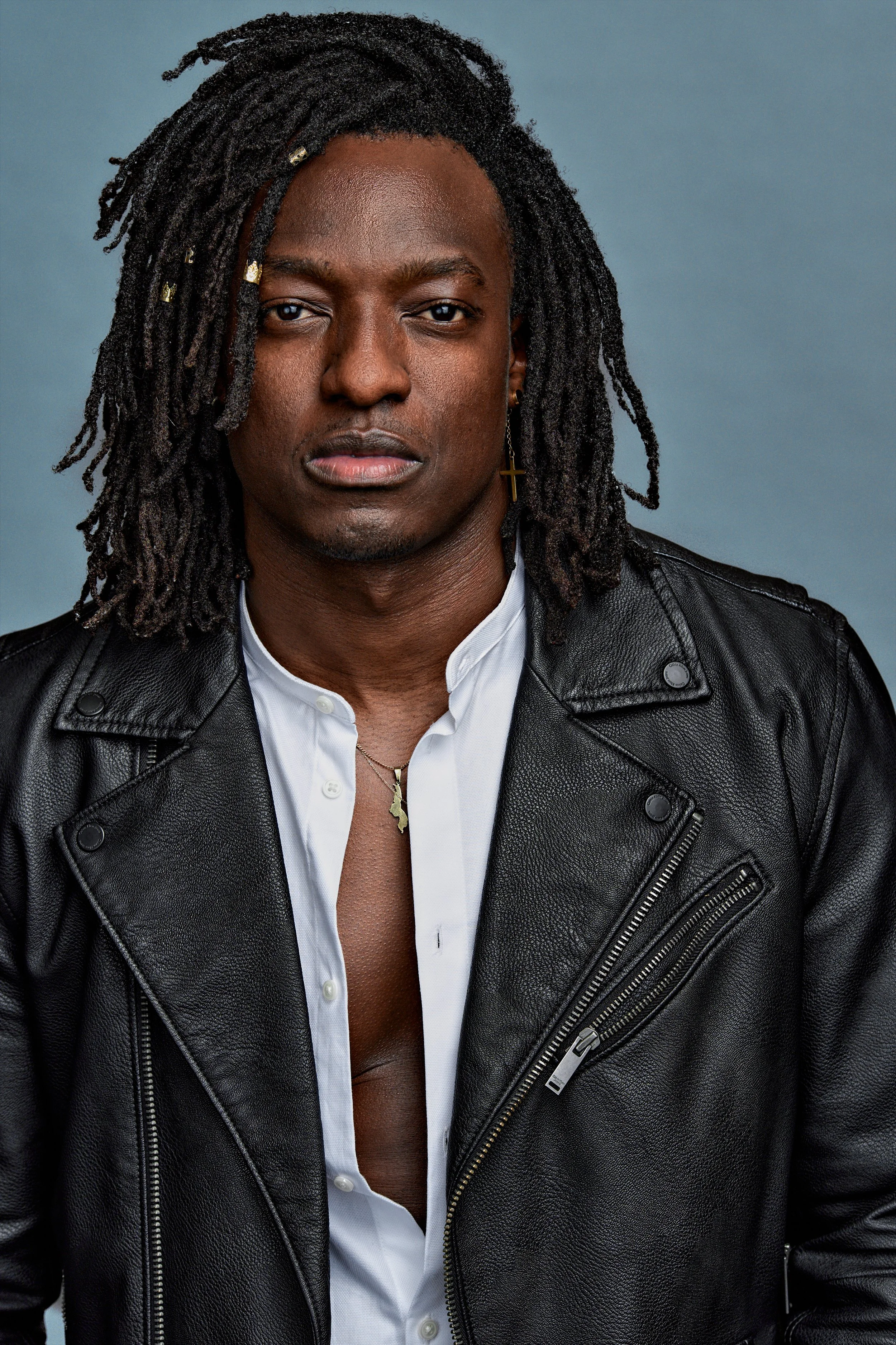Close-up portrait of a Black man with dreadlocks wearing a black leather jacket over a white shirt, with earrings and a necklace, standing against a plain blue background.