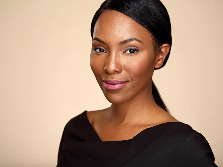 A woman with dark hair pulled back, wearing a black top, smiling softly against a neutral background.