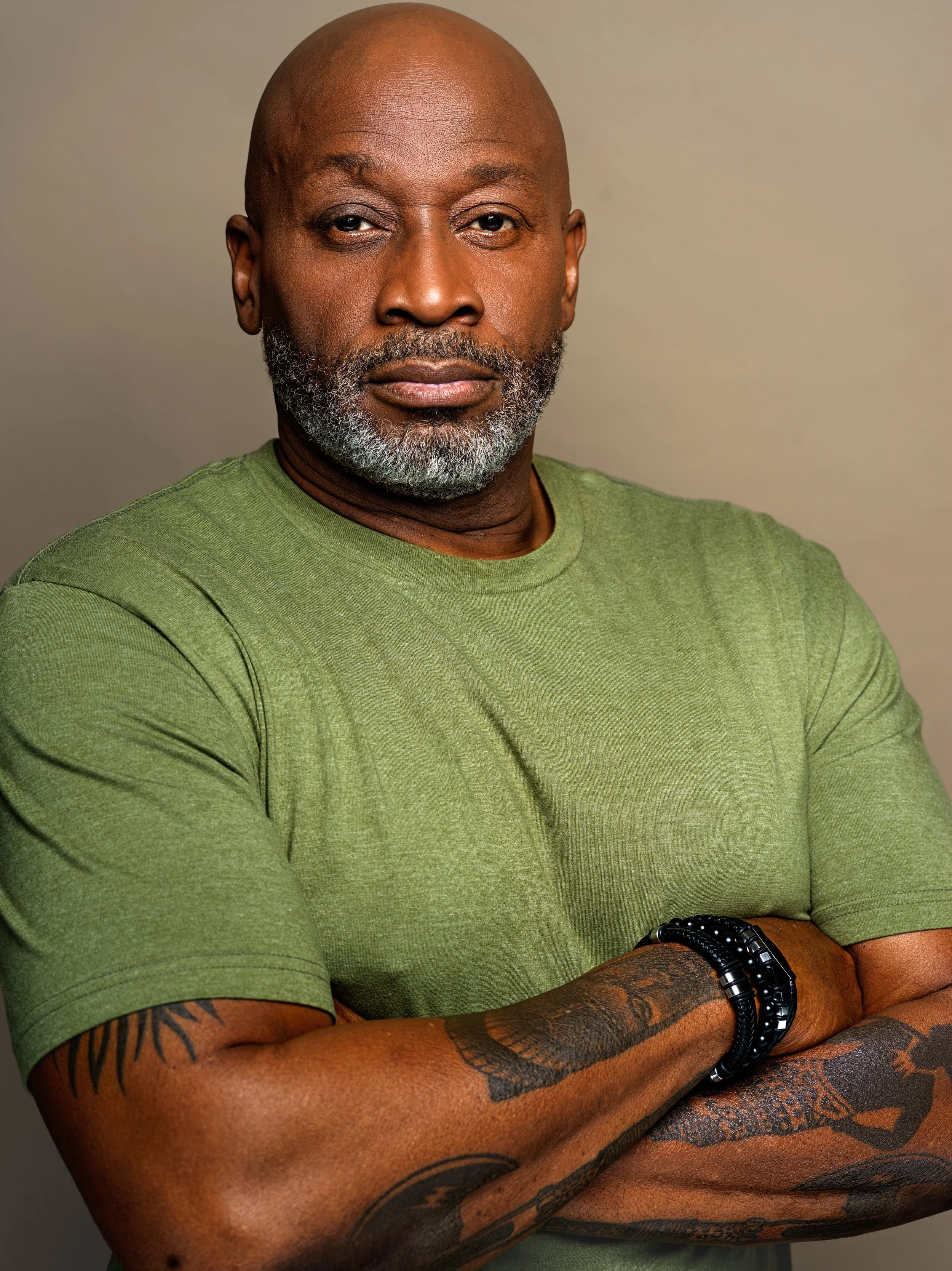 A close-up of a confident man with a bald head and a gray beard, wearing a green T-shirt, black bracelets, and displaying tattoos on his arms, crossing his arms against a plain neutral background.