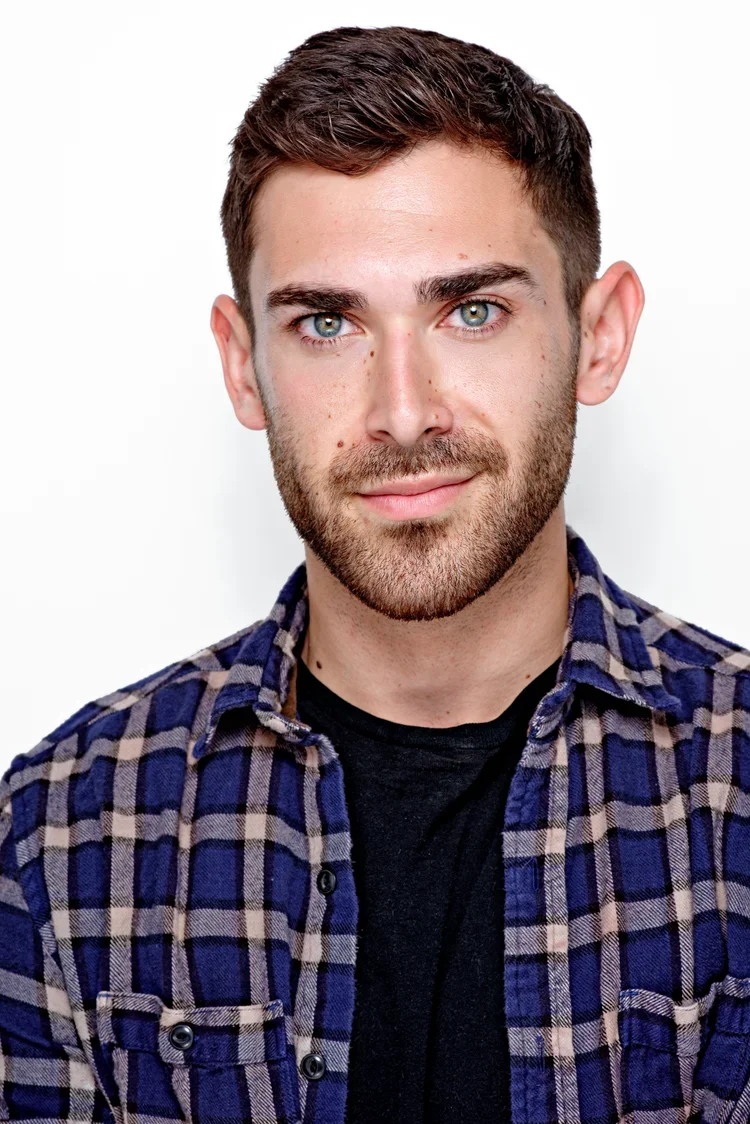 Portrait of a young man with short brown hair, blue eyes, and facial hair, wearing a black t-shirt and a blue plaid shirt against a white background.