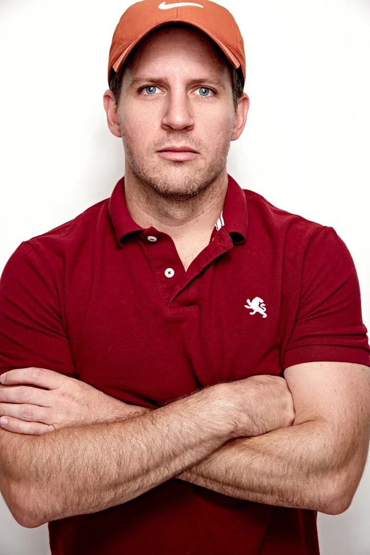 A man with blue eyes and light facial hair wearing a red Nike cap and a red polo shirt with an embroidered lion logo, standing with arms crossed against a plain white background.