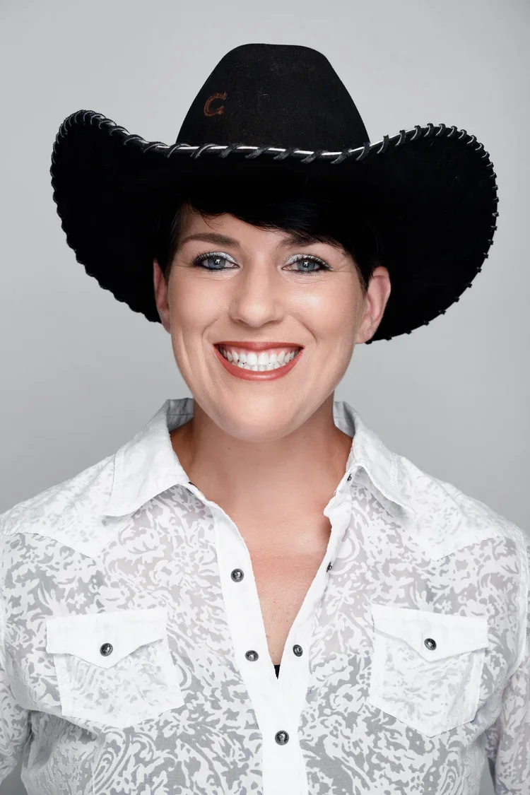 A woman smiling wearing a black cowboy hat and a white patterned button-up shirt against a light gray background.