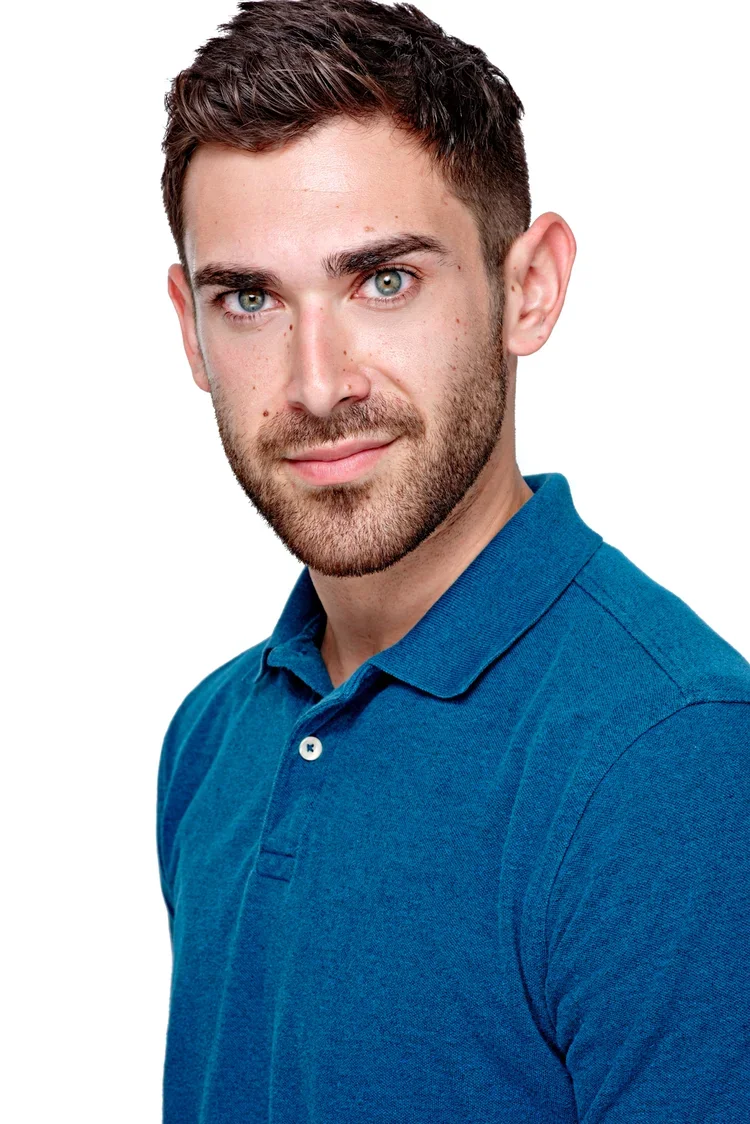 A portrait of a young man with short brown hair and a beard, wearing a blue polo shirt, looking at the camera against a white background.
