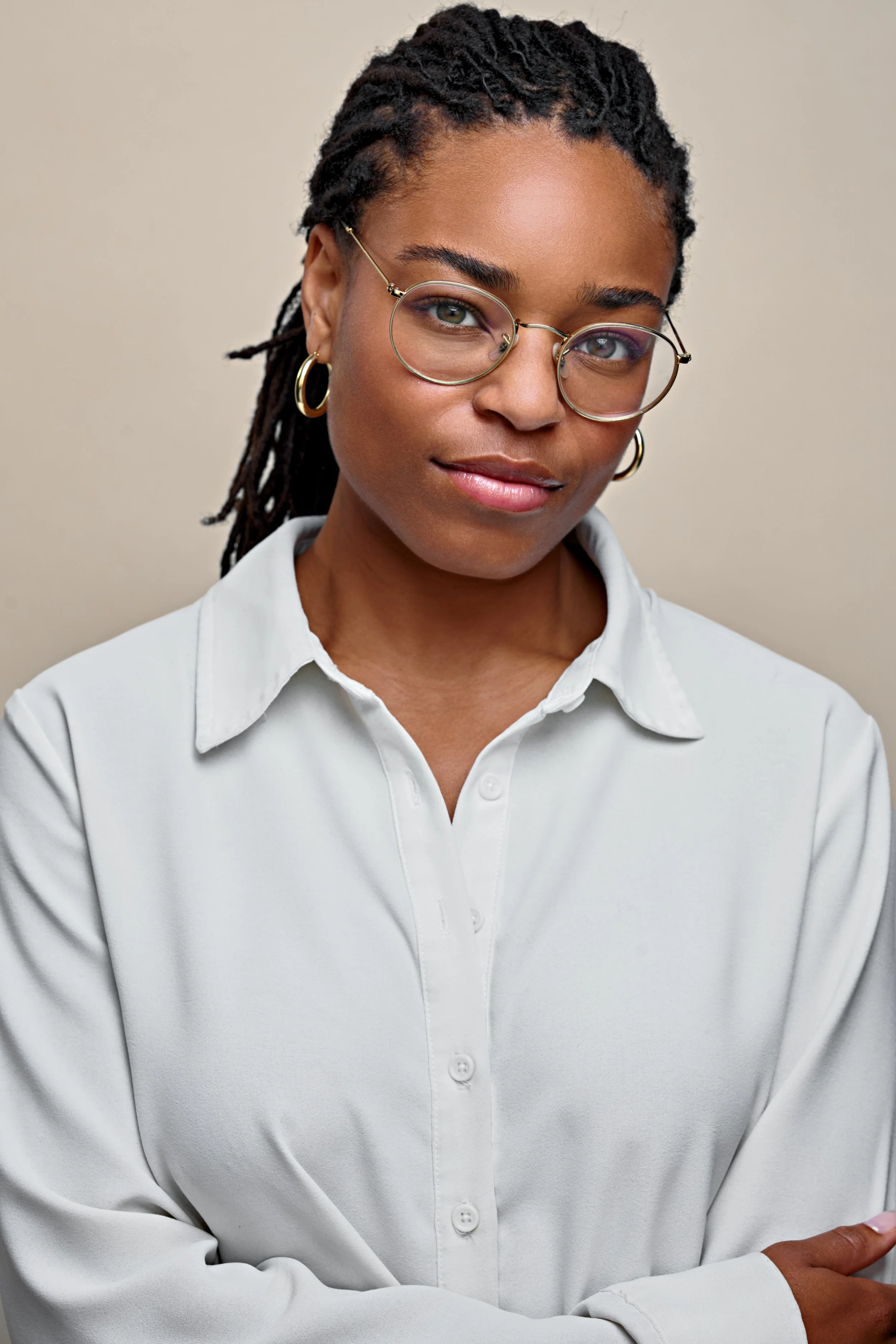 A woman with short dreadlocks wearing glasses and gold hoop earrings, dressed in a white collared shirt, crosses her arms and looks at the camera with a slight smile against a beige background.