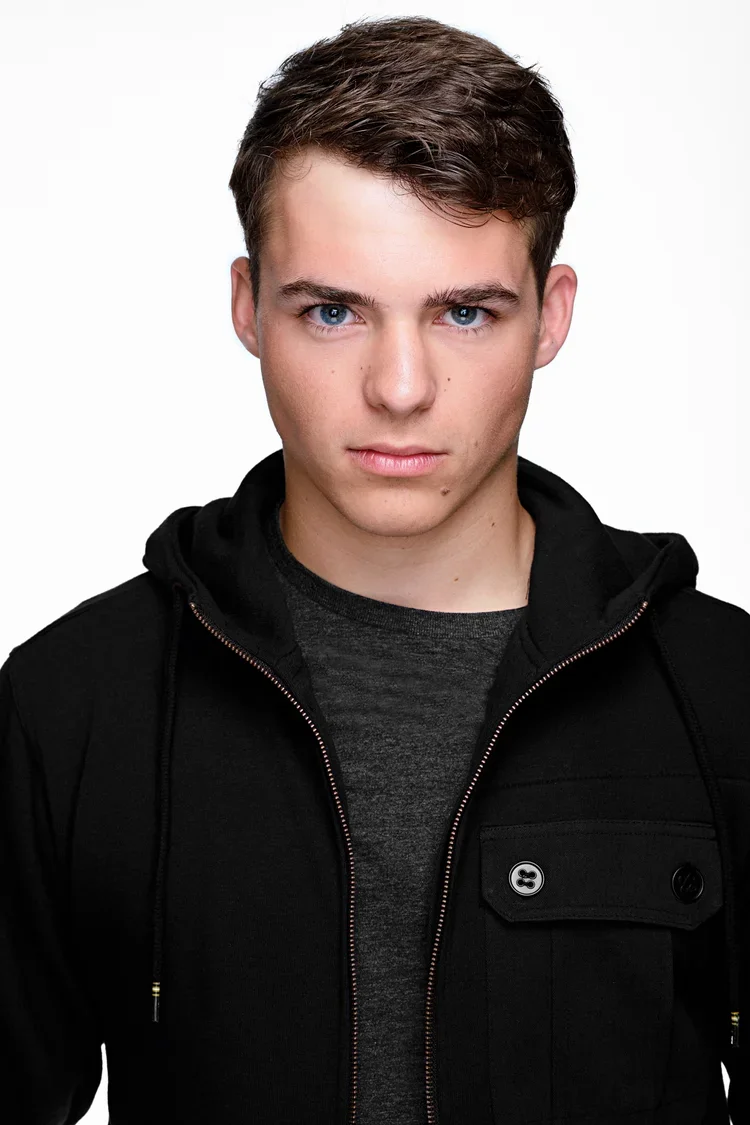 A young man with short brown hair and blue eyes looking directly at the camera, wearing a black hoodie over a dark gray shirt against a white background.