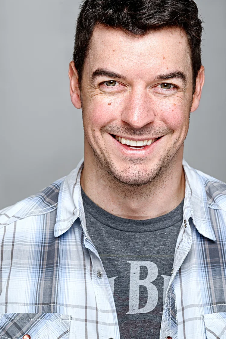 A smiling man with short dark hair, light skin, wearing a plaid shirt over a dark T-shirt with a letter 'B' on it, against a gray background.