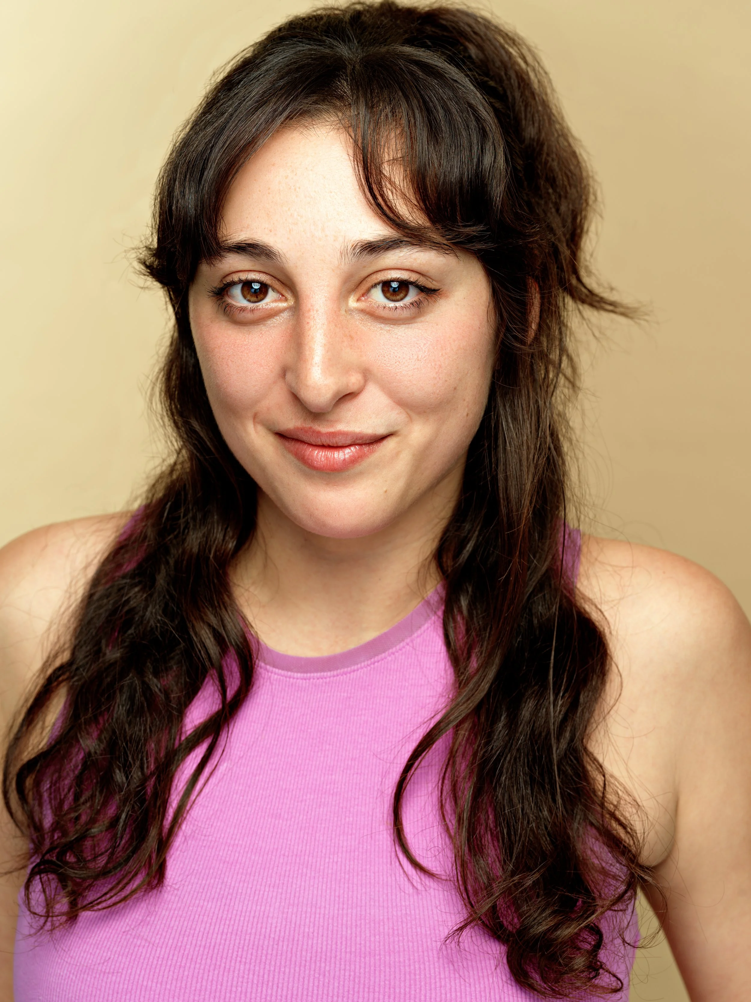 A young woman with long, dark wavy hair wearing a pink sleeveless top, smiling with a neutral beige background.