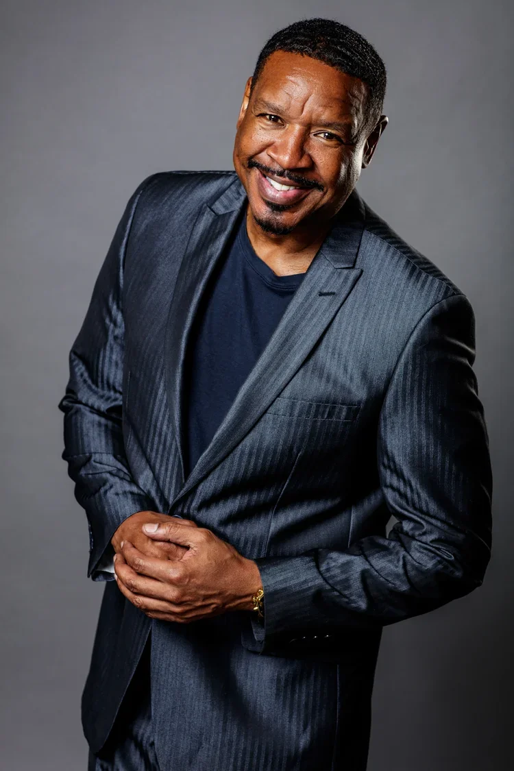 A smiling man in a dark suit and black t-shirt, posing against a gray background.