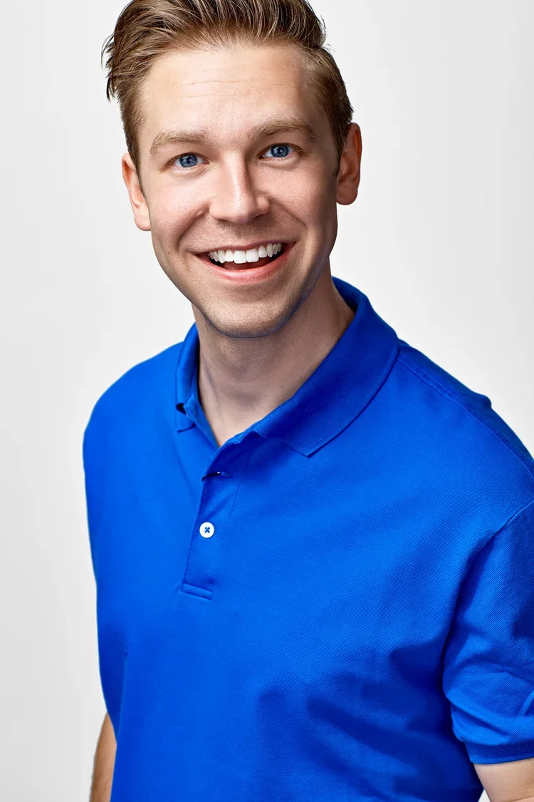 Young man with blue eyes and light brown hair smiling, wearing a blue polo shirt, against a plain white background.