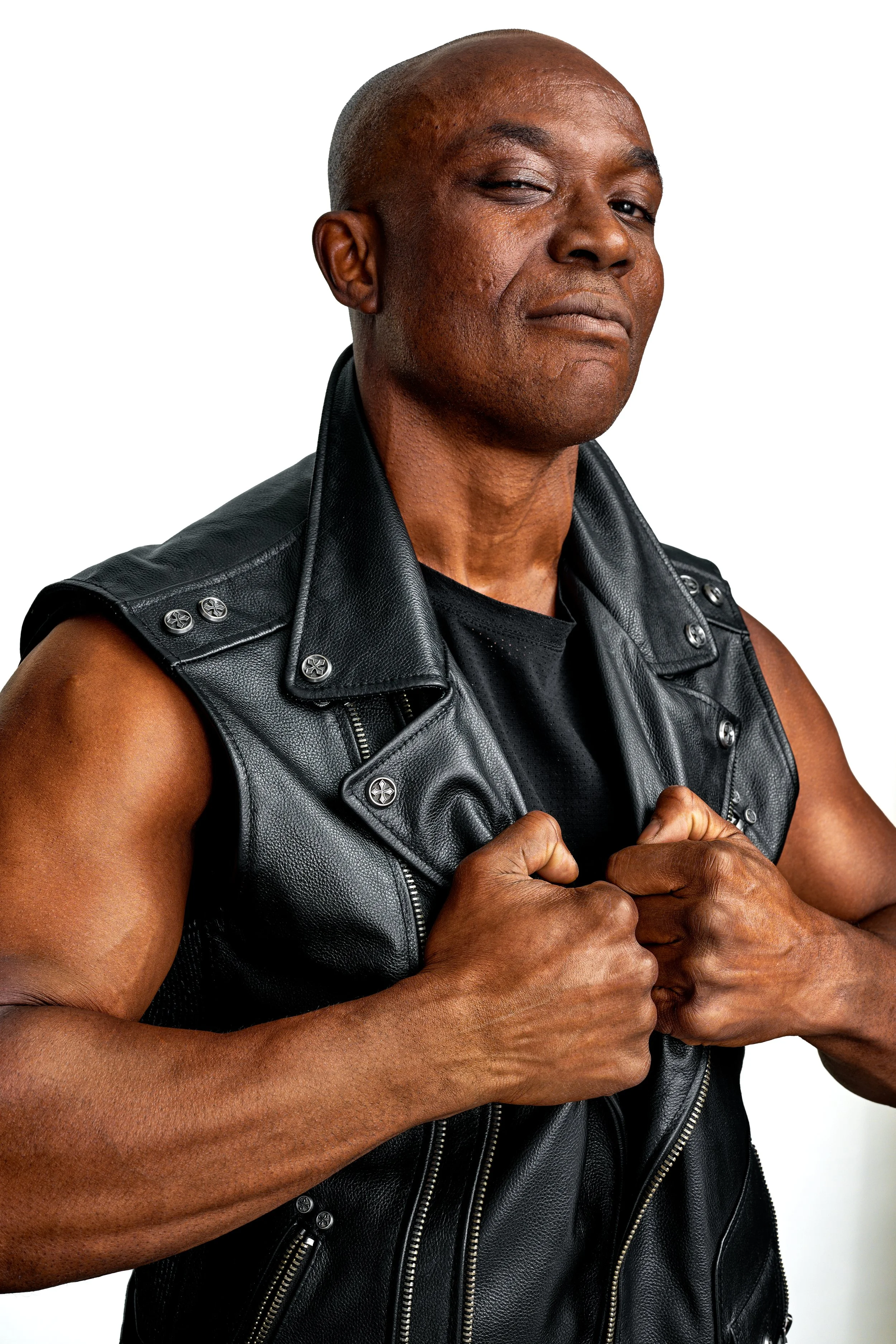 A confident man with a shaved head, wearing a black leather vest, posing with fists clenched.