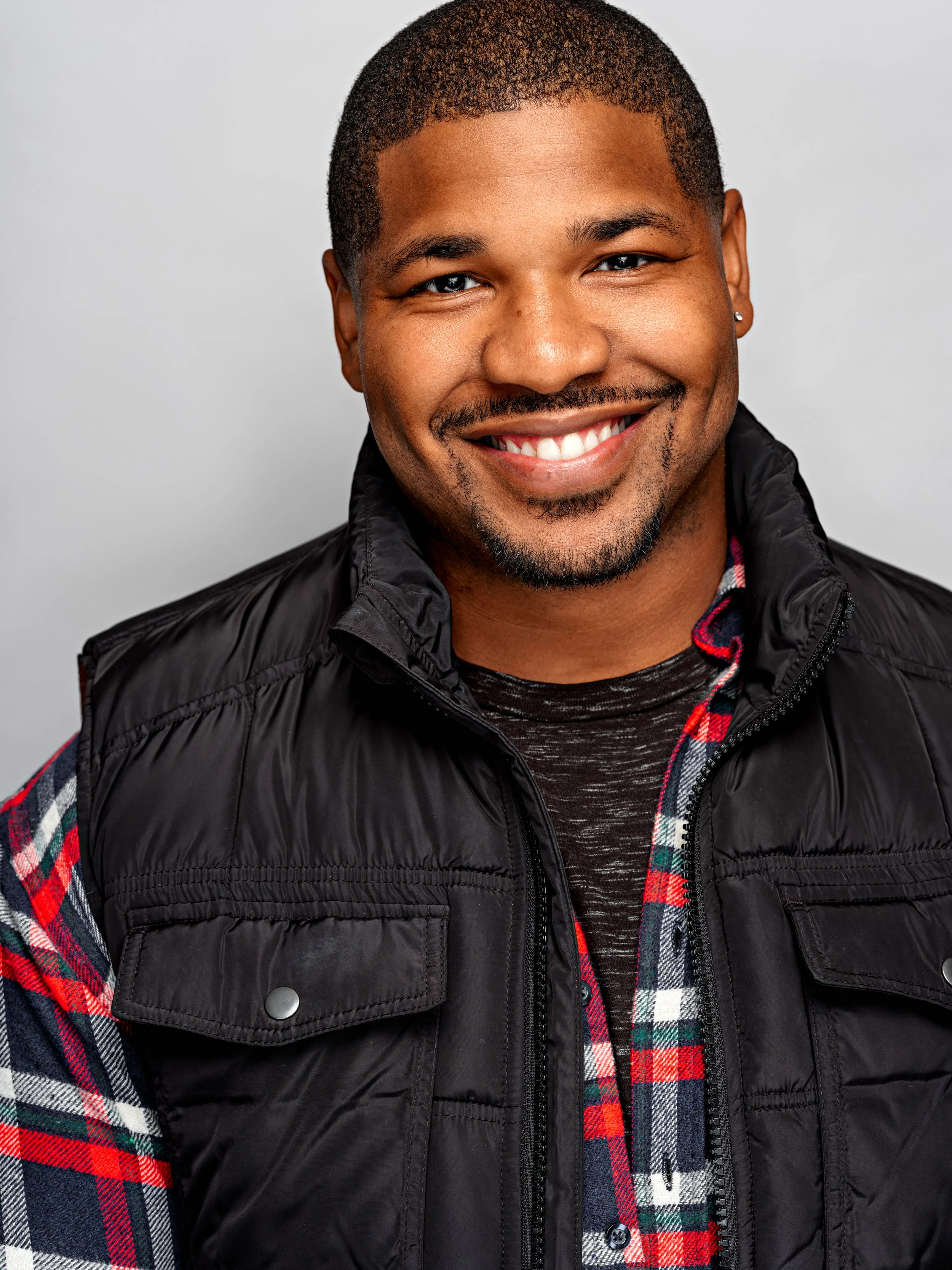 Portrait of a smiling man with short hair, a goatee, wearing a black puffer vest over a red and green plaid shirt, with a gray background.