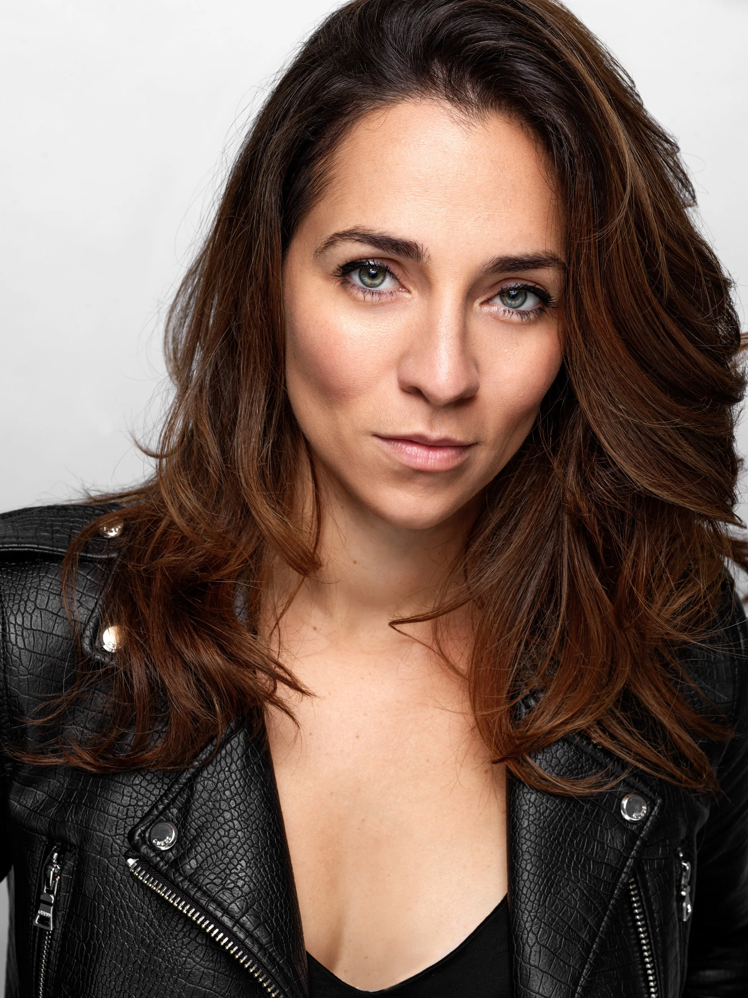 Close-up portrait of a woman with wavy brown hair, green eyes, wearing a black leather jacket with silver zippers and buttons.