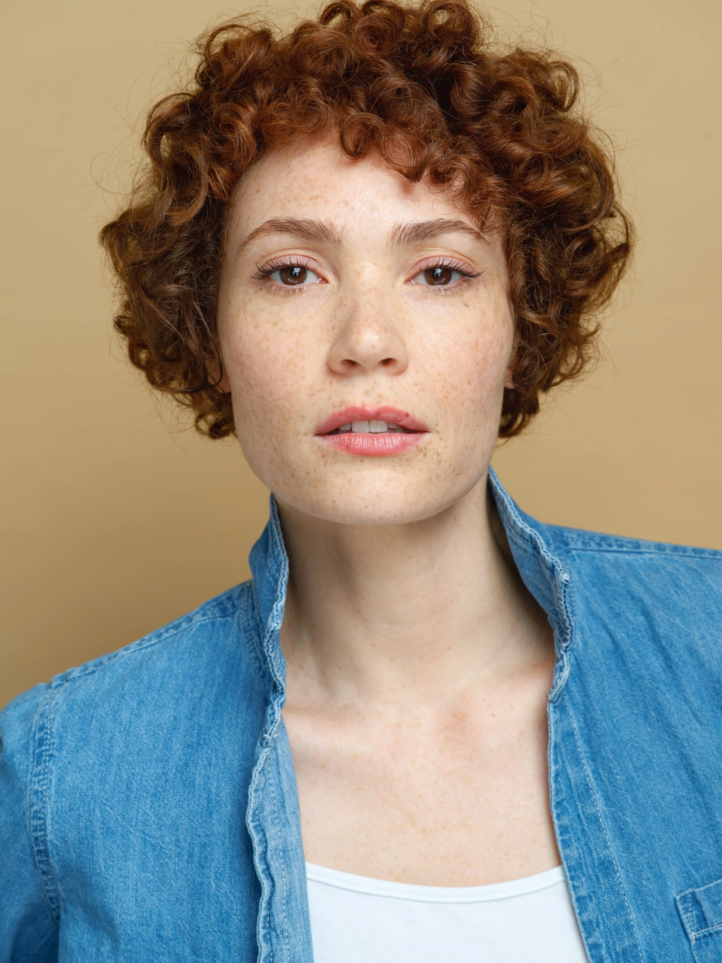 Close-up portrait of a woman with red curly hair, freckles, wearing a blue denim jacket over a white top, standing against a beige background.