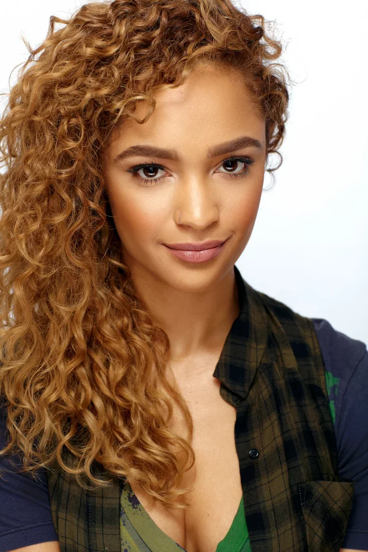 Close-up of a young woman with curly red hair, wearing a gray and black plaid shirt layered over a green top, looking at the camera with a subtle smile.