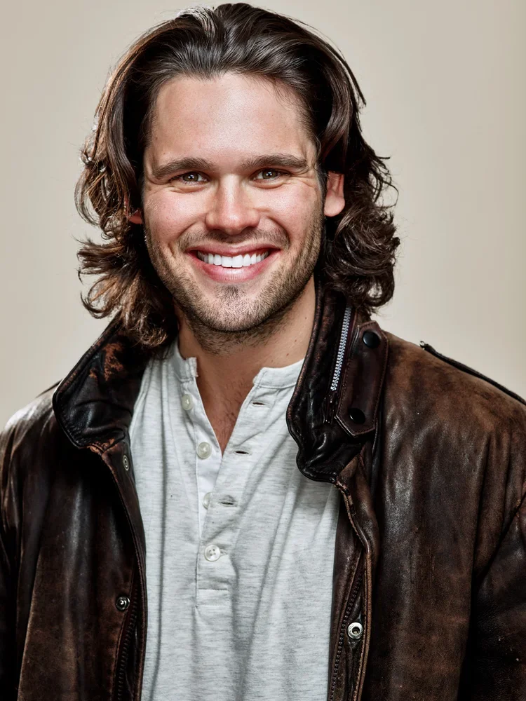 A young man with long, wavy brown hair, smiling, wearing a white button-up shirt and a brown leather jacket.