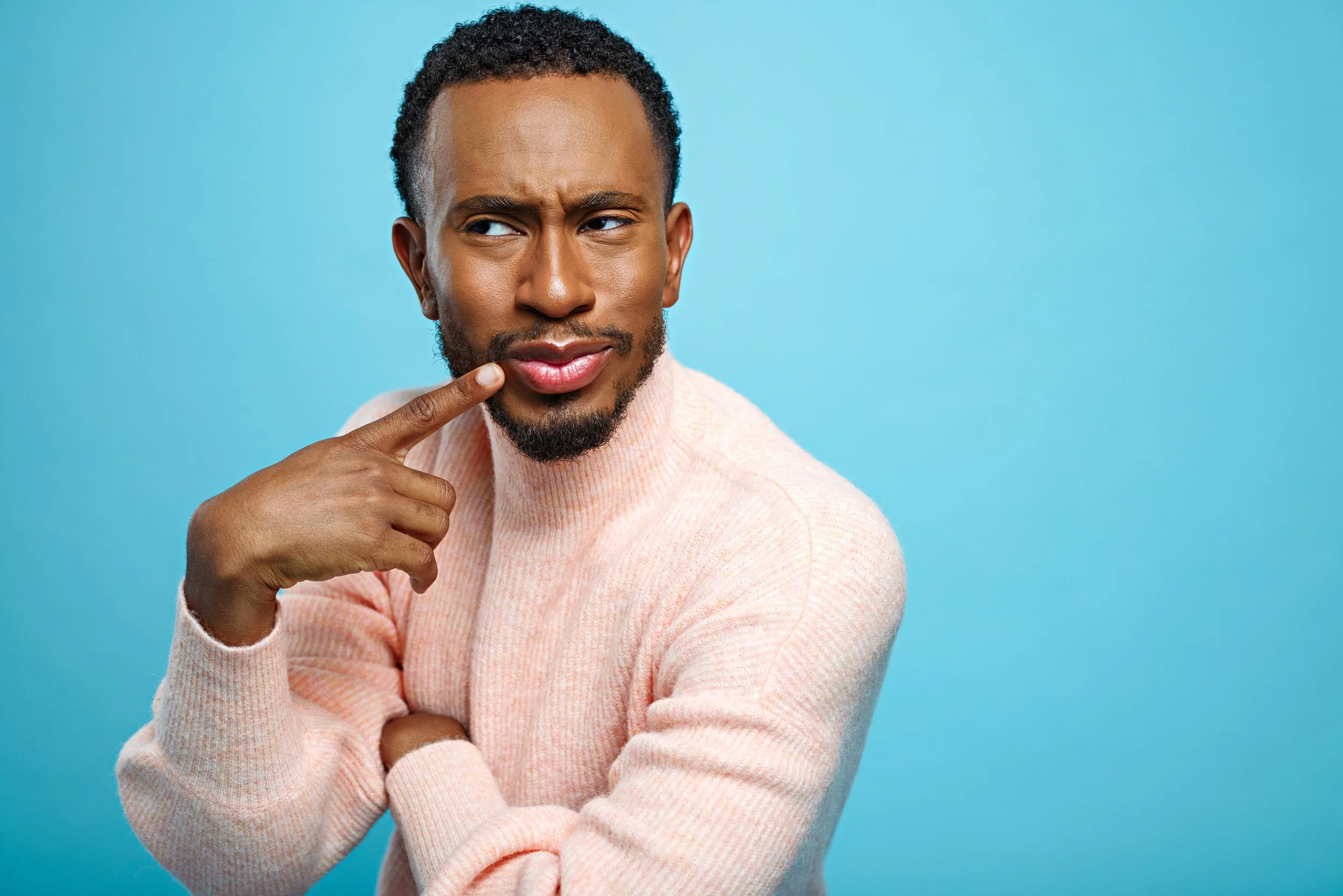 A man with dark curly hair and a beard is touching his cheek and looking to the side with a puzzled expression, wearing a light pink sweater, against a bright blue background.