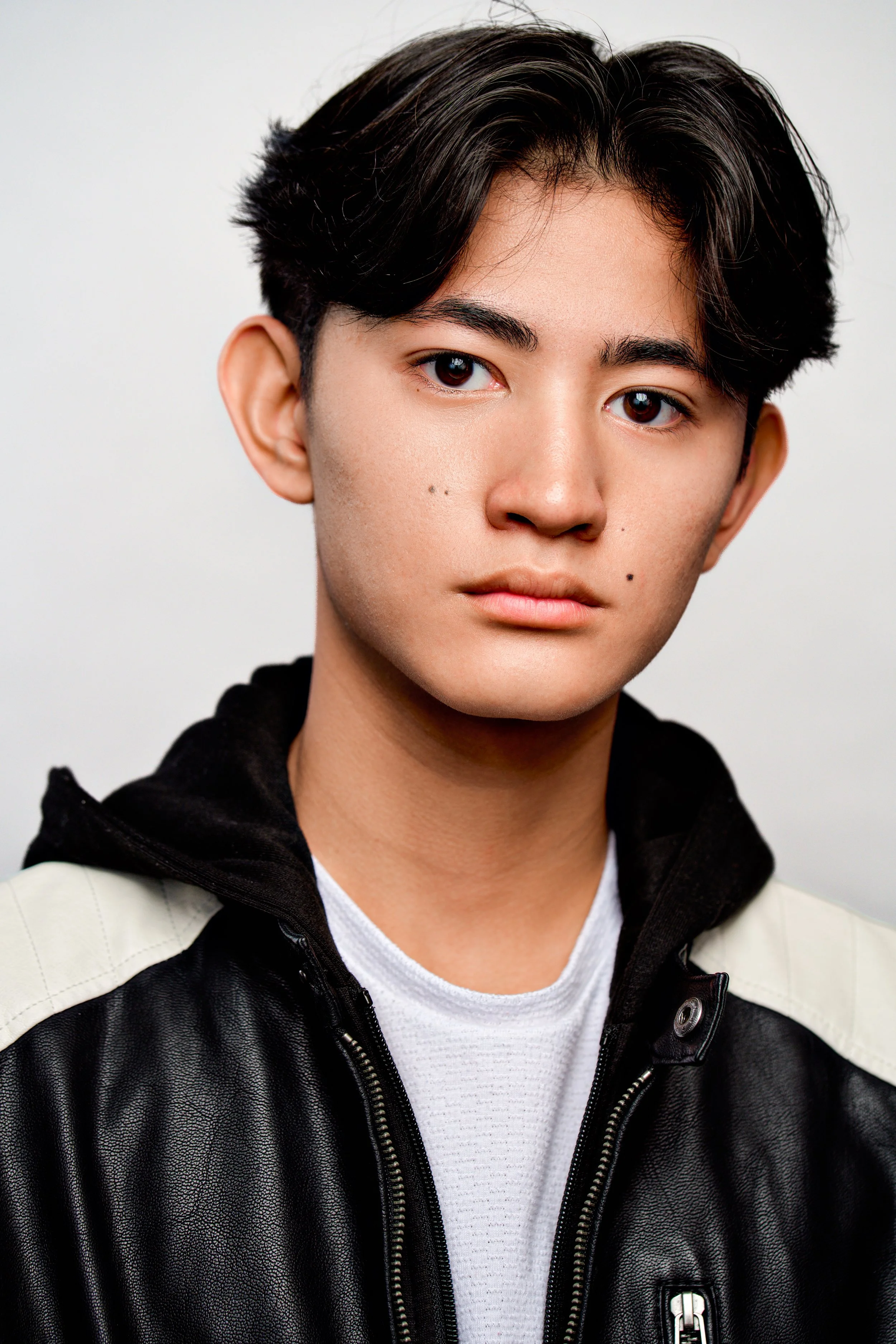Close-up of a young person with short black hair, wearing a black and beige leather jacket and white shirt, looking directly at the camera against a plain light background.
