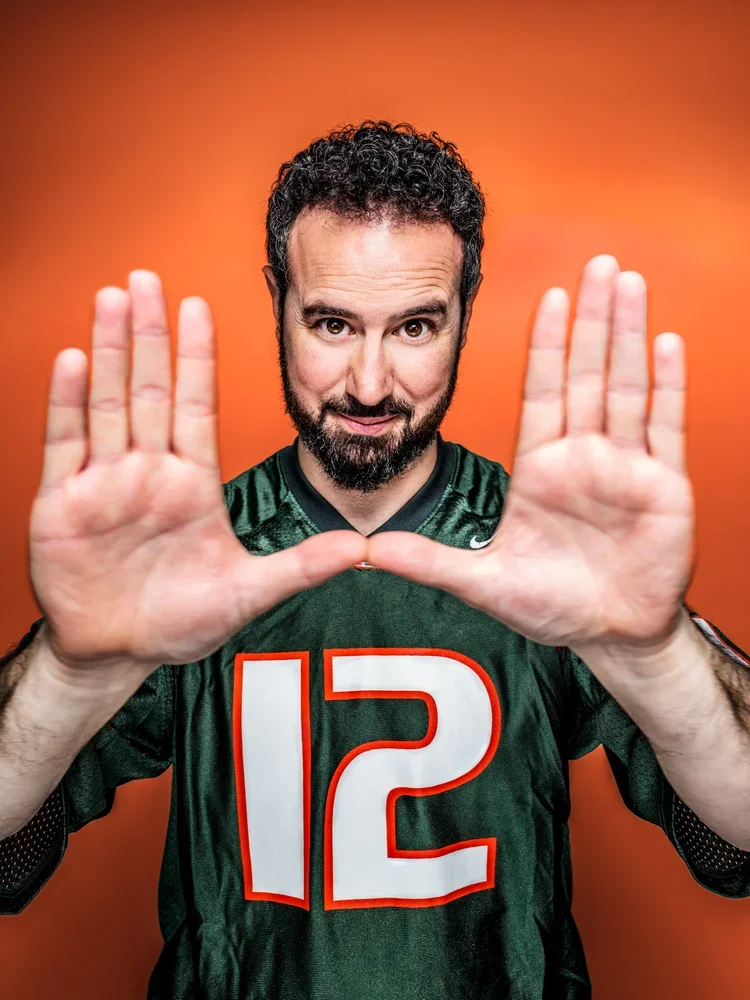 Man in a green sports jersey with the number 12, making a framing gesture with his hands, against an orange background.