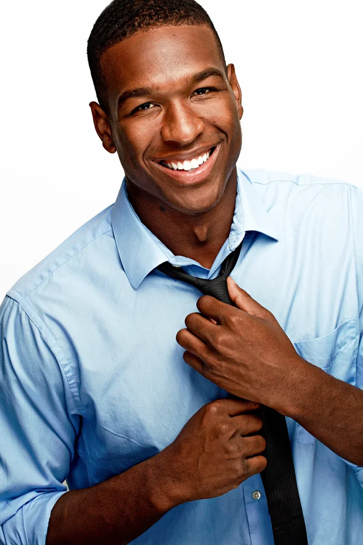 Smiling young man in a light blue dress shirt, adjusting a black tie against a plain white background.