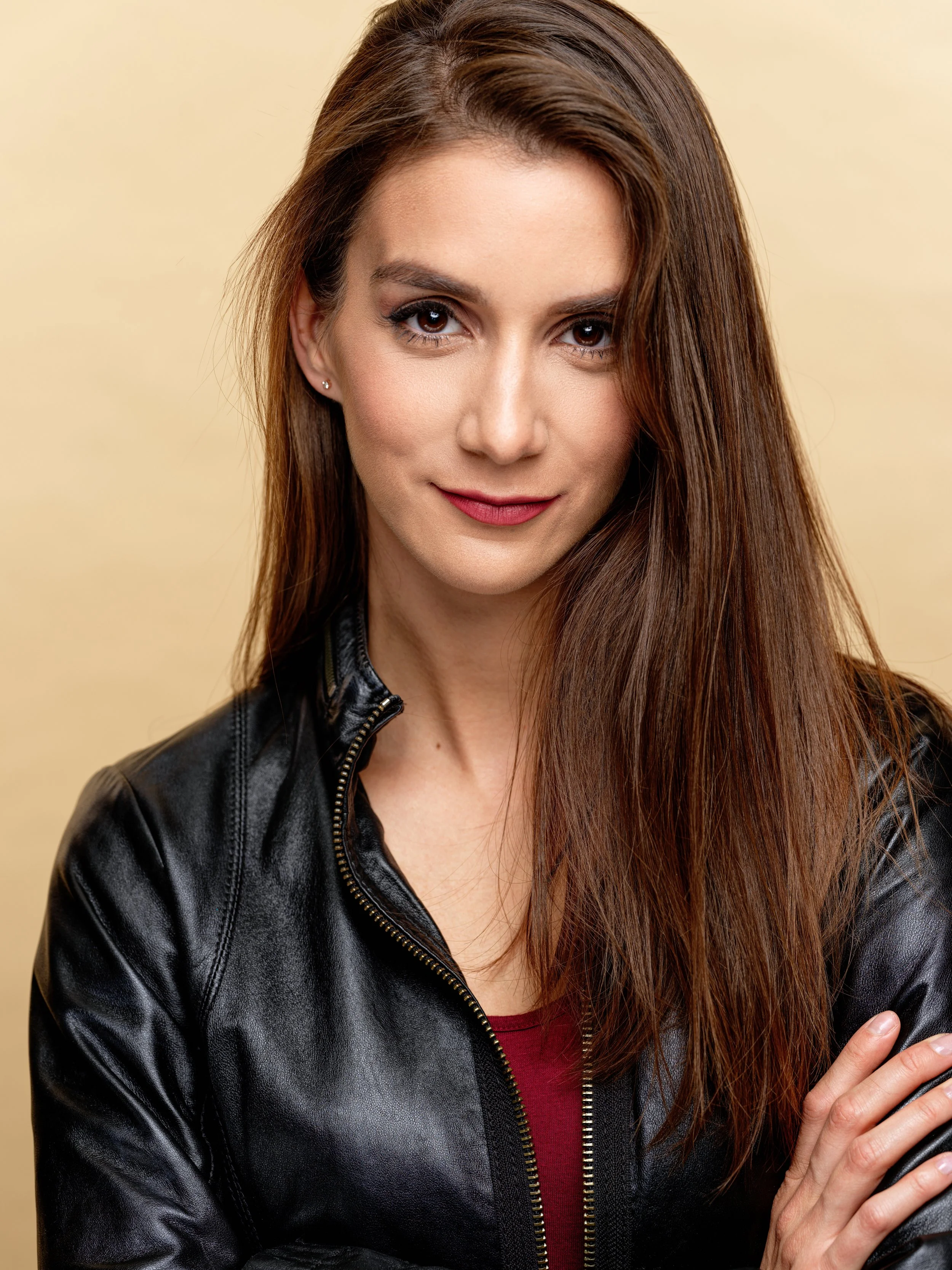 A woman with long brown hair, brown eyes, and light makeup, wearing a black leather jacket and a red top, standing with her arms crossed against a plain beige background.