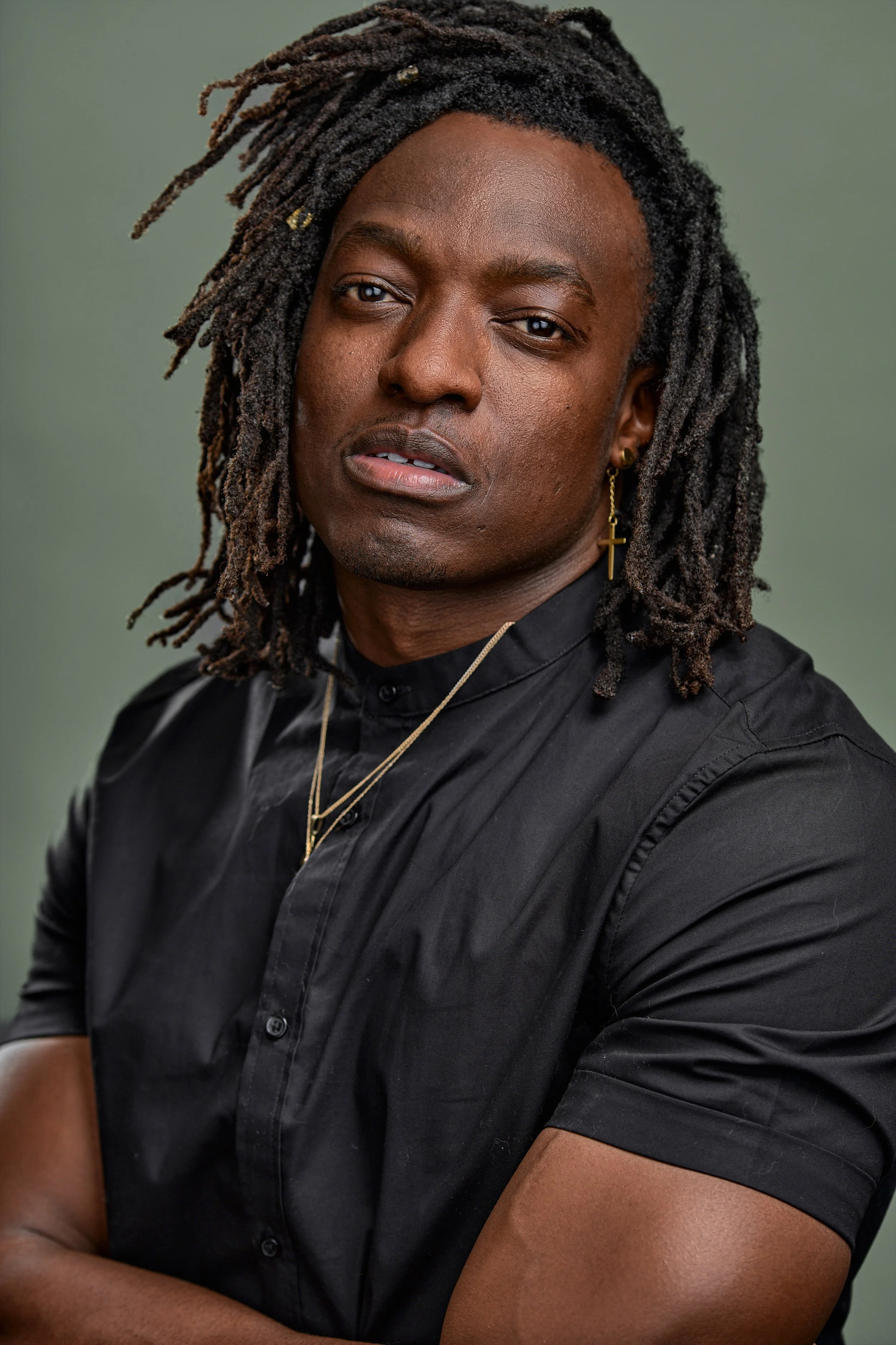 Close-up of a person with long dreadlocks, wearing a black shirt, cross earring, and layered necklaces, with a neutral background.