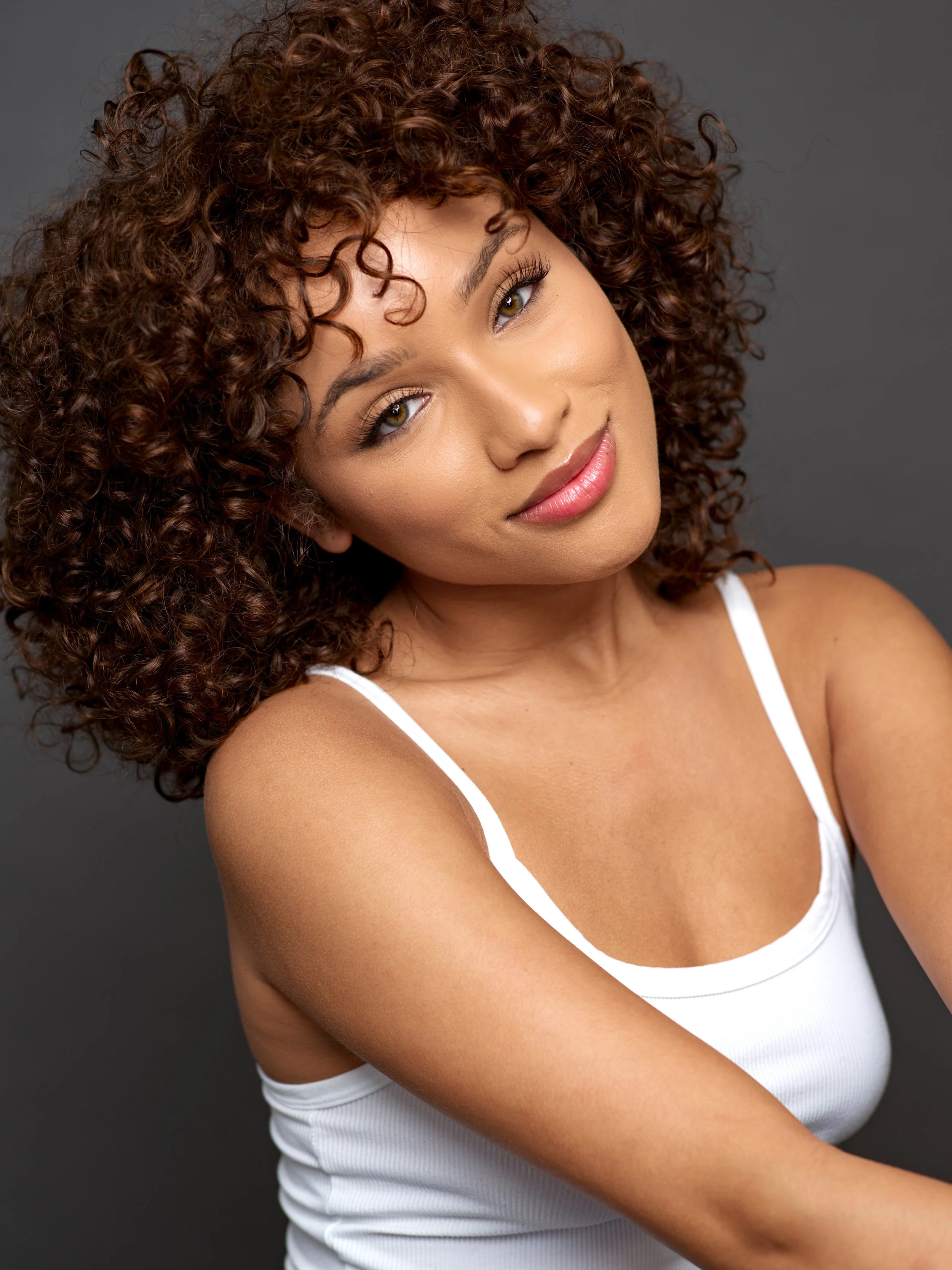 Close-up portrait of a woman with curly brown hair, wearing a white tank top, smiling slightly, against a gray background.