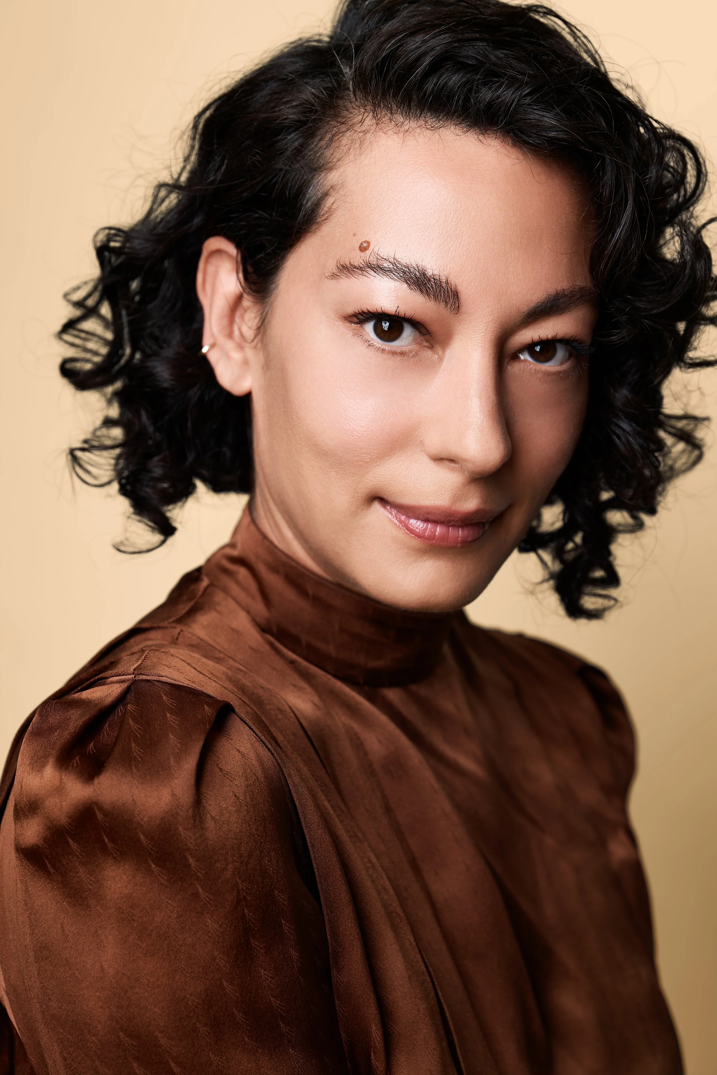 A woman with dark, curly hair and light skin, wearing a brown silk blouse with a subtle pattern, looking directly at the camera with a slight smile.