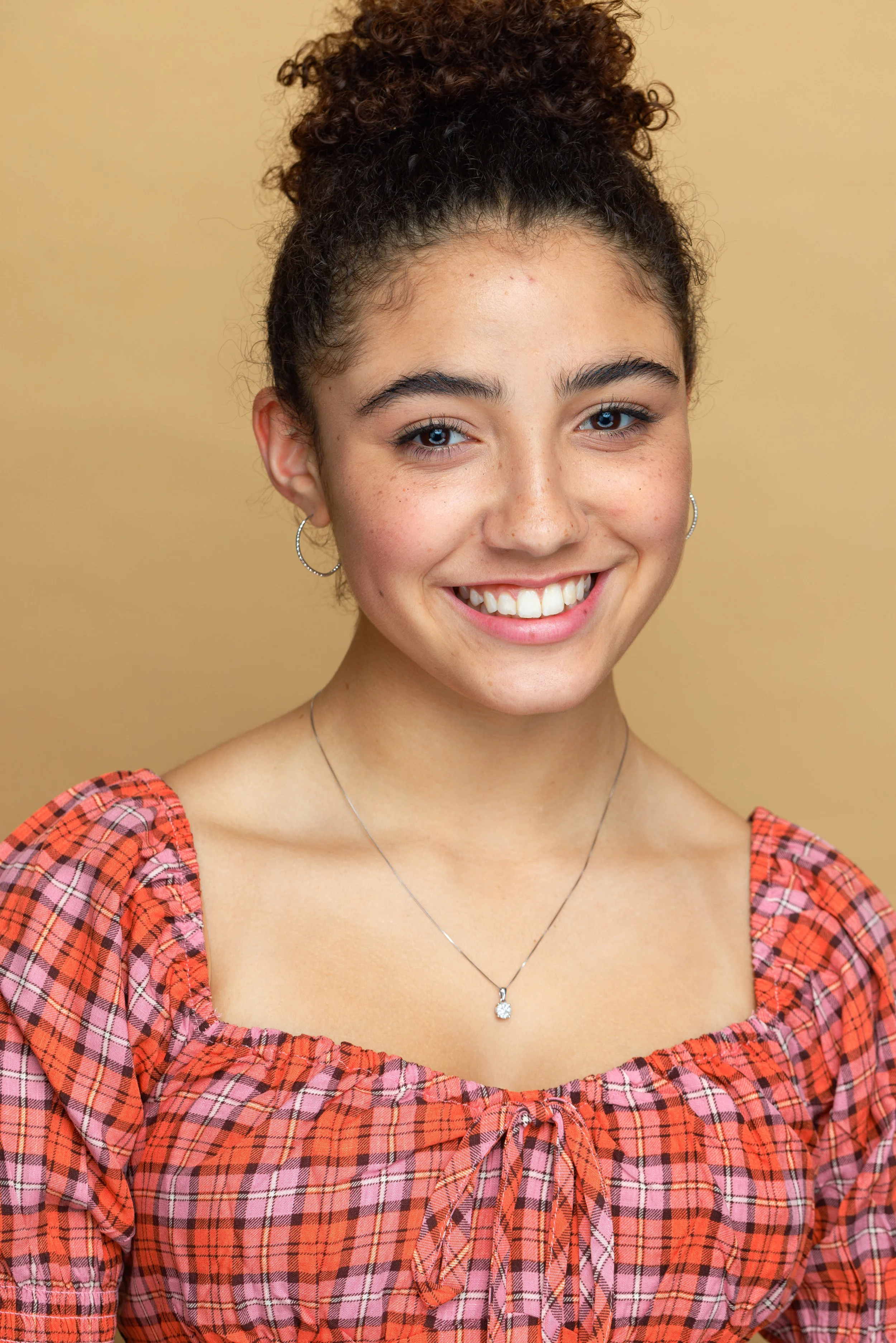 A young woman with curly brown hair in a bun, smiling, wearing silver hoop earrings, a silver necklace with a pendant, and a red plaid dress with a bow at the chest, standing against a beige background.