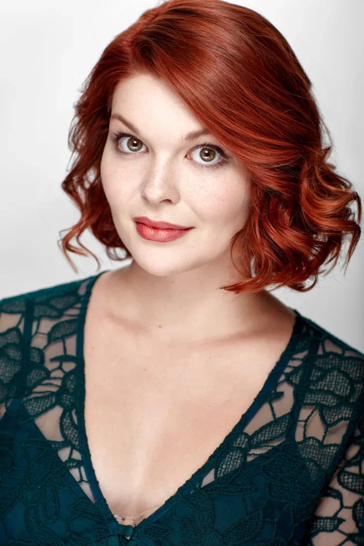 A woman with red, wavy hair and fair skin wearing a dark green lace top.