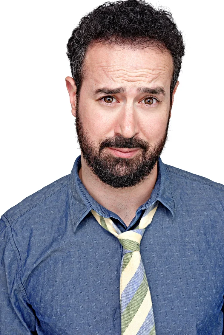 A man with curly dark hair and a beard, wearing a blue button-up shirt and a striped tie, making a confused or skeptical facial expression.