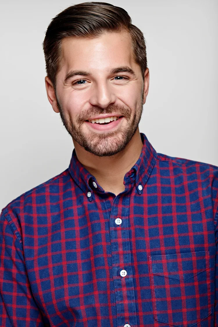 A young man with a beard and short brown hair smiling, wearing a blue and red checkered button-up shirt, against a plain light gray background.