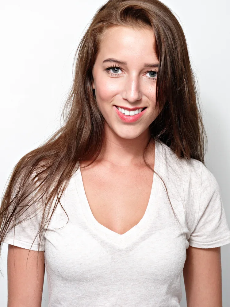 A young woman with long brown hair and blue eyes smiling, wearing a white V-neck T-shirt, against a plain light background.