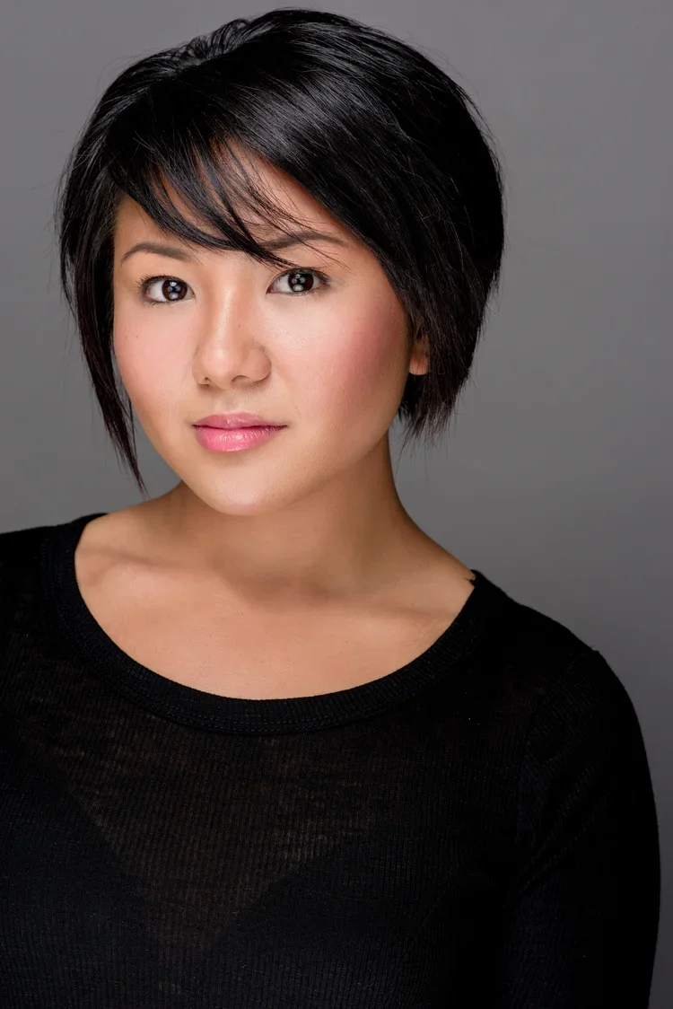 A portrait of a young Asian woman with short, layered black hair, wearing a black top, looking directly at the camera with a neutral expression against a gray background.