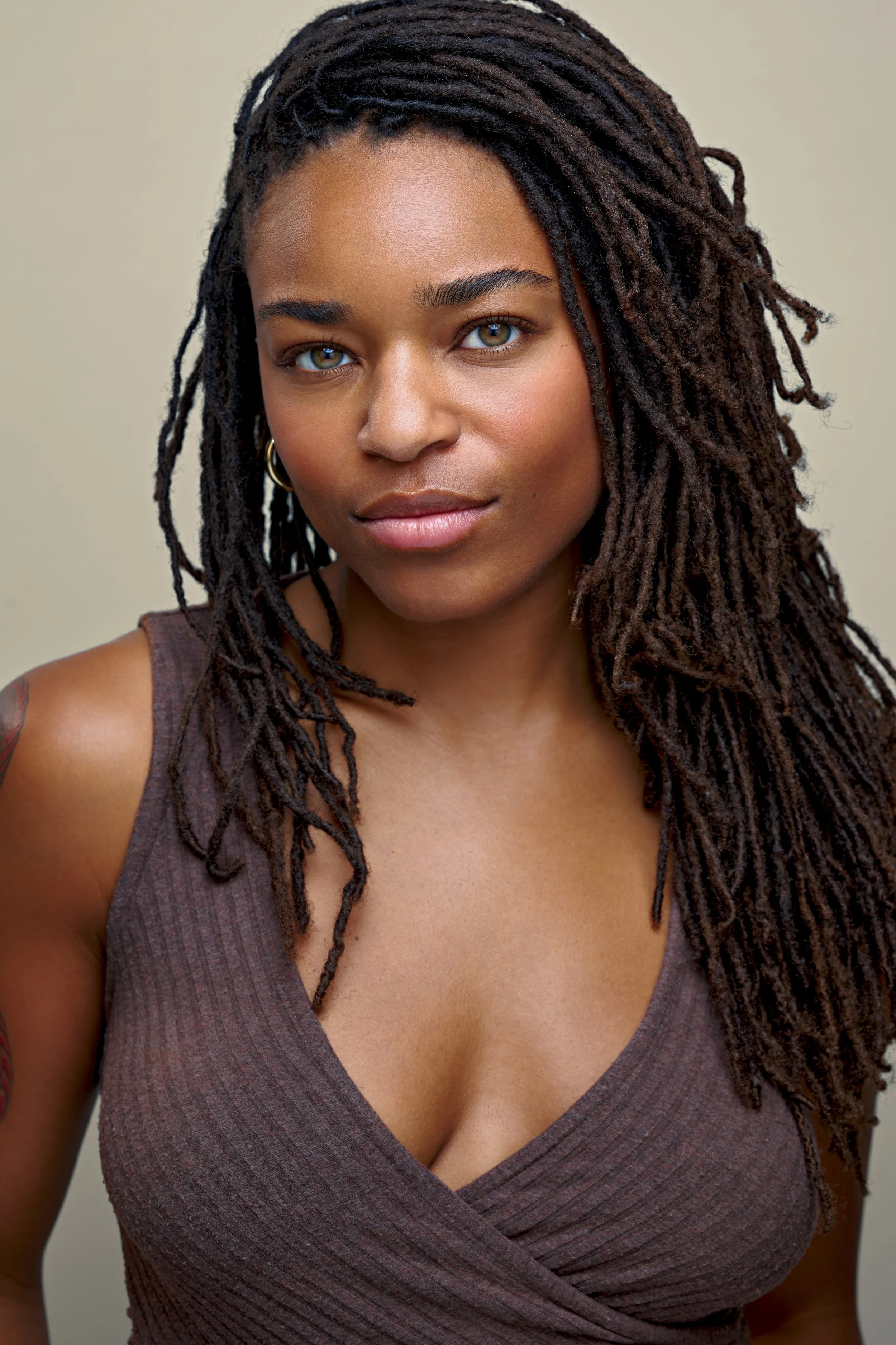 Close-up portrait of a woman with medium brown skin, dark dreadlocks, and green eyes, wearing a sleeveless brown top and small hoop earrings, looking confidently at the camera.