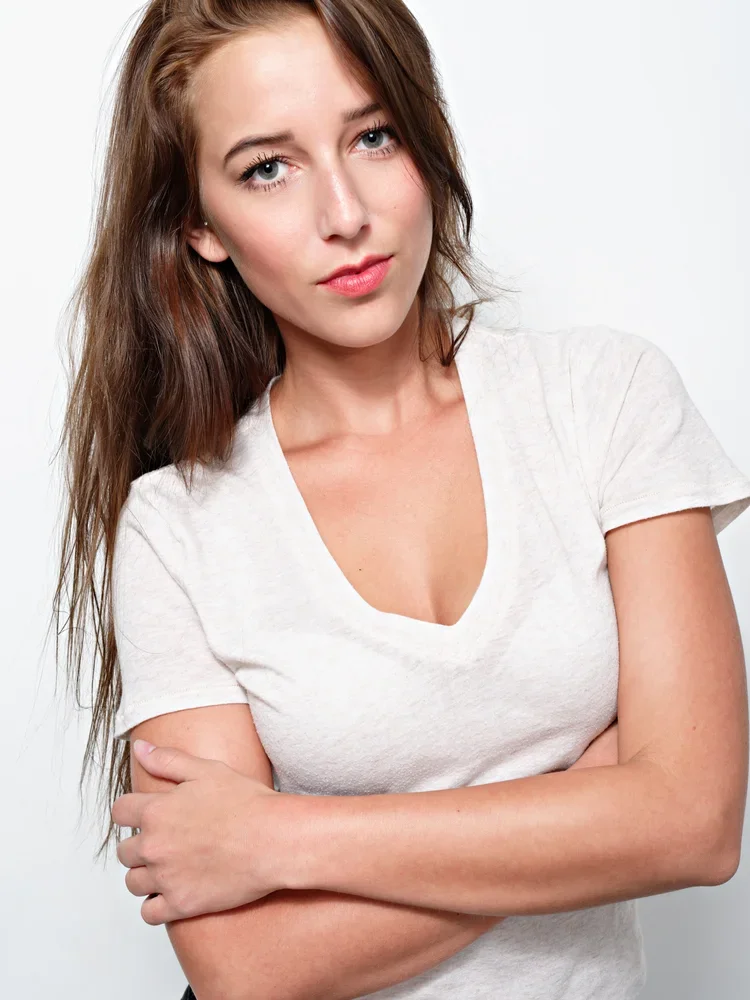 A young woman with long brown hair and blue eyes wearing a white t-shirt, standing with arms crossed, looking at the camera with a neutral expression.