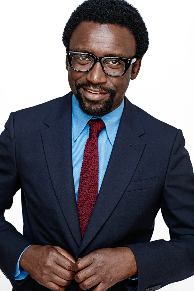 A man in a navy suit, light blue shirt, and red tie, wearing black glasses, smiling at the camera against a white background.