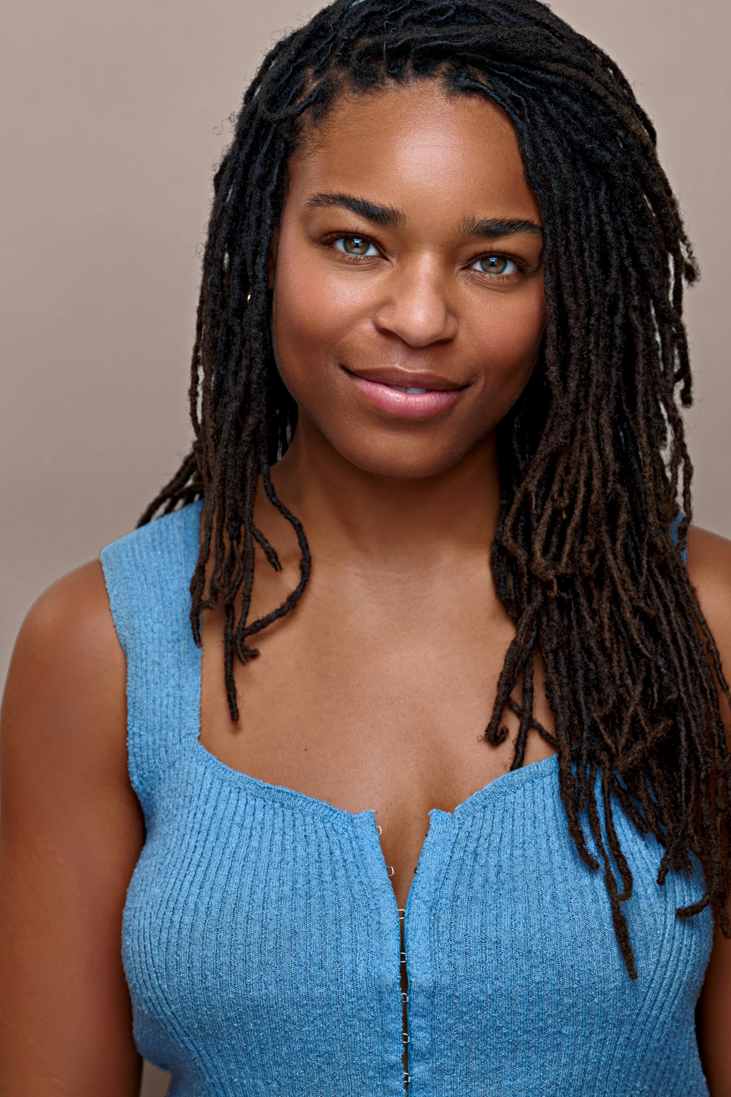 A young woman with dark, curly, shoulder-length hair, wearing a sleeveless blue top with a ribbed texture and a zipper down the front, smiling against a plain, light-colored background.