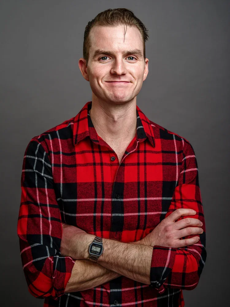 A young man with short, slicked-back hair and fair skin, standing against a gray background. He's smiling softly and has his arms crossed, wearing a red and black plaid button-up shirt and a digital wristwatch.