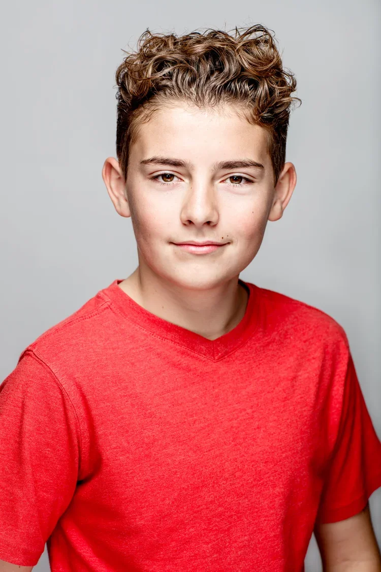 A teenage boy with curly light brown hair wearing a red t-shirt, smiling slightly, against a plain gray background.