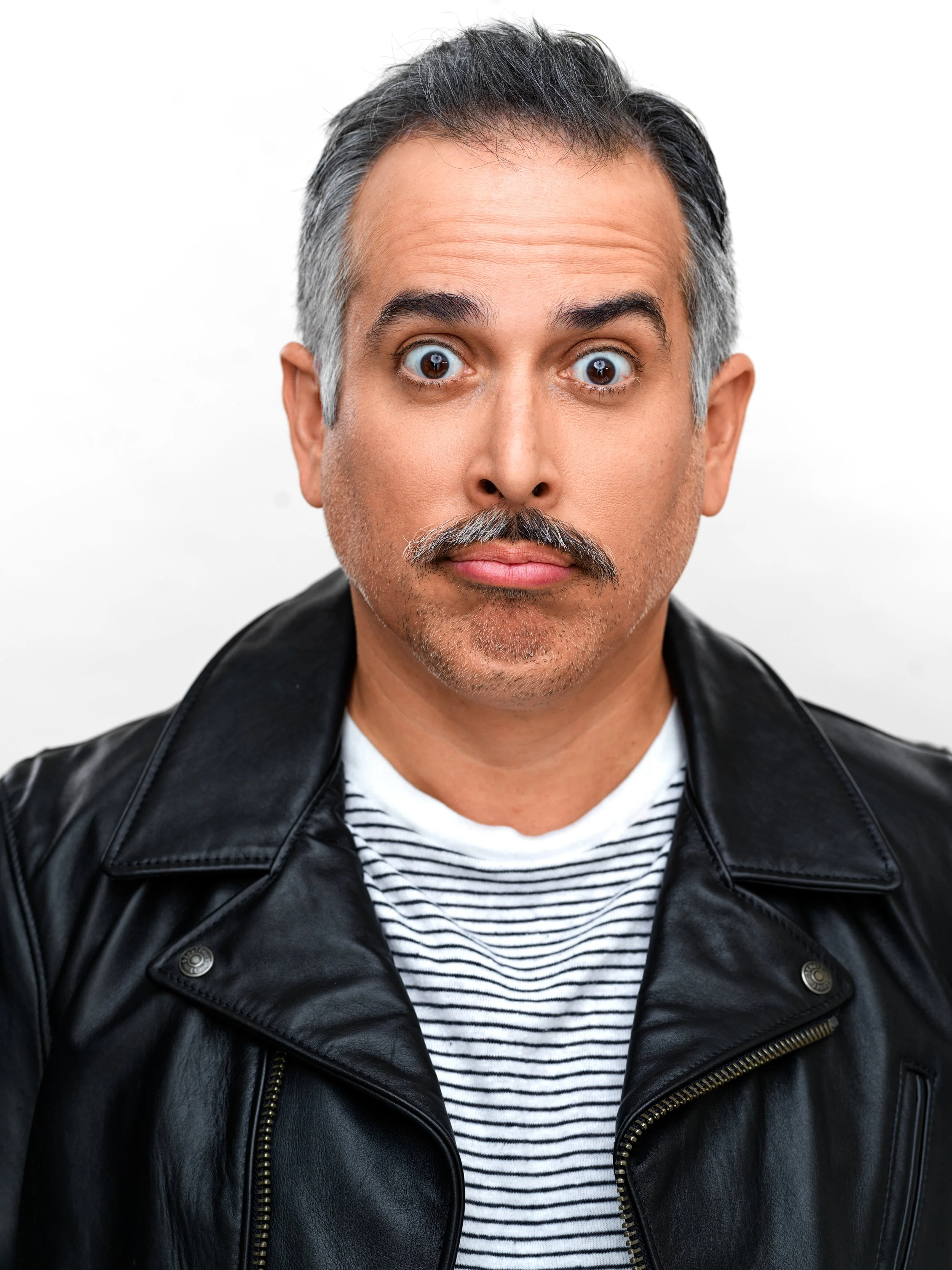 A middle-aged man with salt-and-pepper hair and a mustache, wearing a black leather jacket and a striped shirt, looking directly at the camera with a surprised expression.