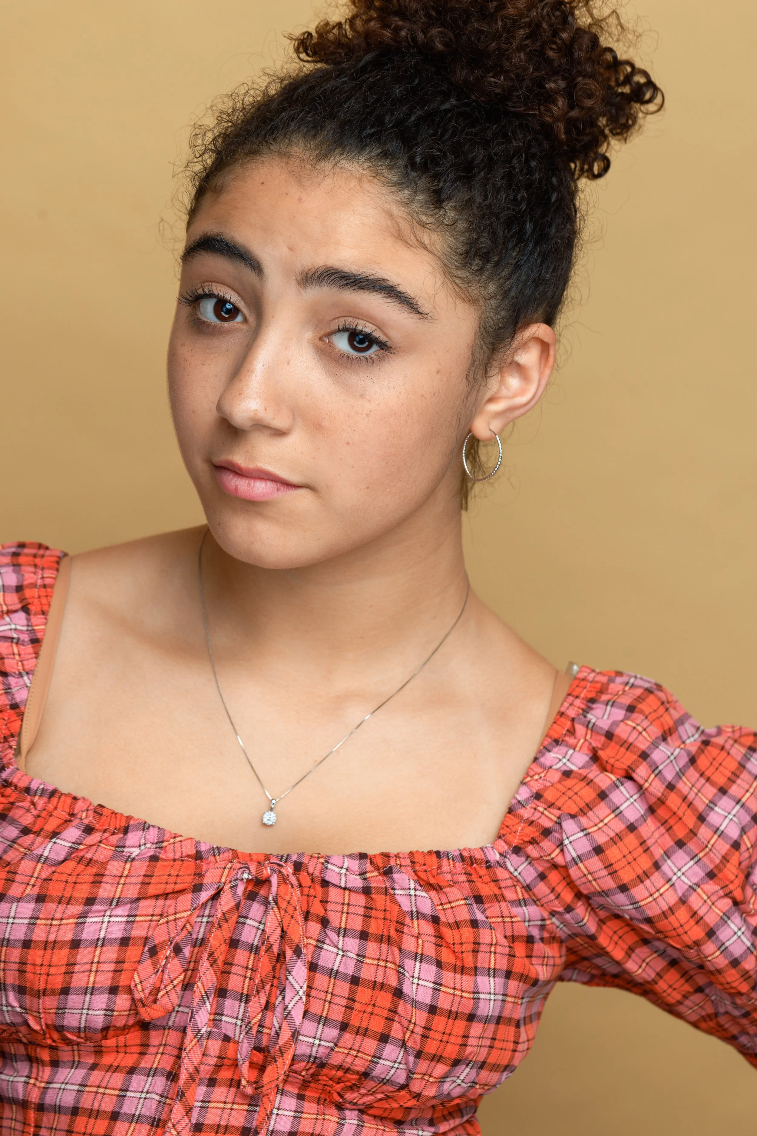 Close-up of a young woman with curly dark hair in a bun wearing hoop earrings and a silver necklace, dressed in a red and black checkered top, against a beige background.