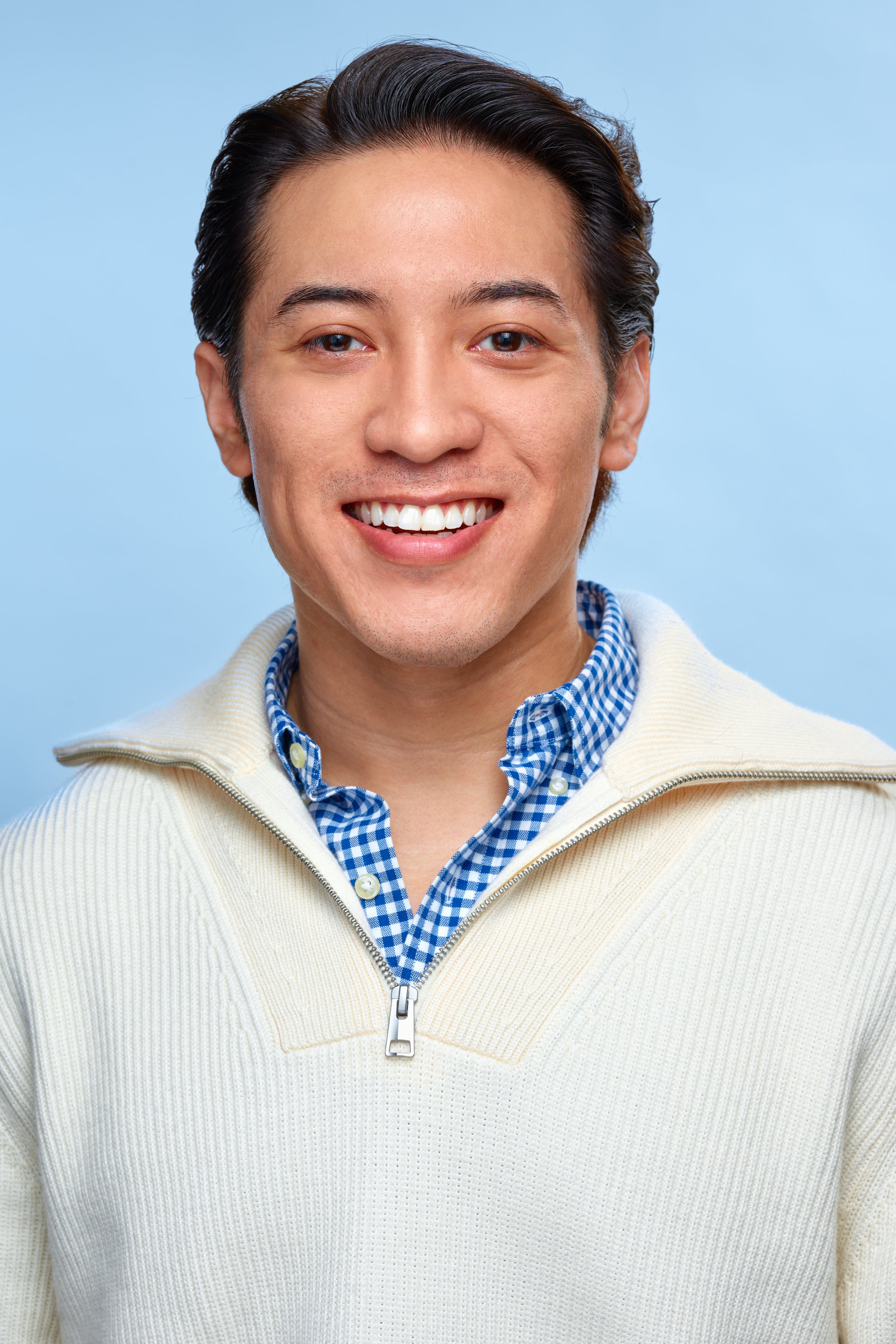 A man with light skin and dark, slicked-back hair smiling, wearing a blue checkered shirt under a cream-colored sweater zip-up front against a light blue background.