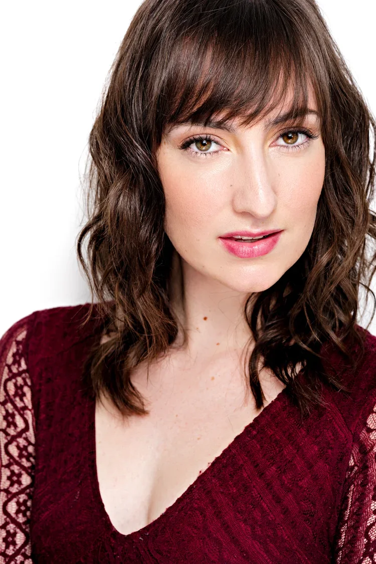 A woman with wavy brown hair and fair skin, wearing a burgundy lace top, looks at the camera with a slight smile, against a plain white background.