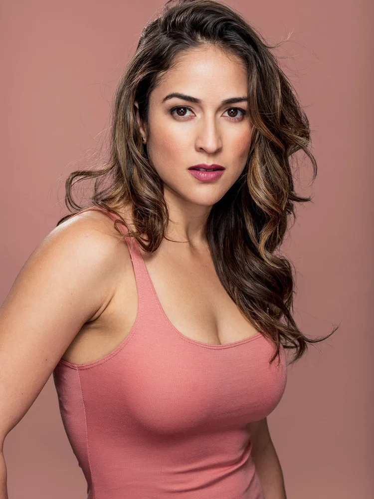 A woman with wavy brown hair in a pink tank top posing against a pink background.